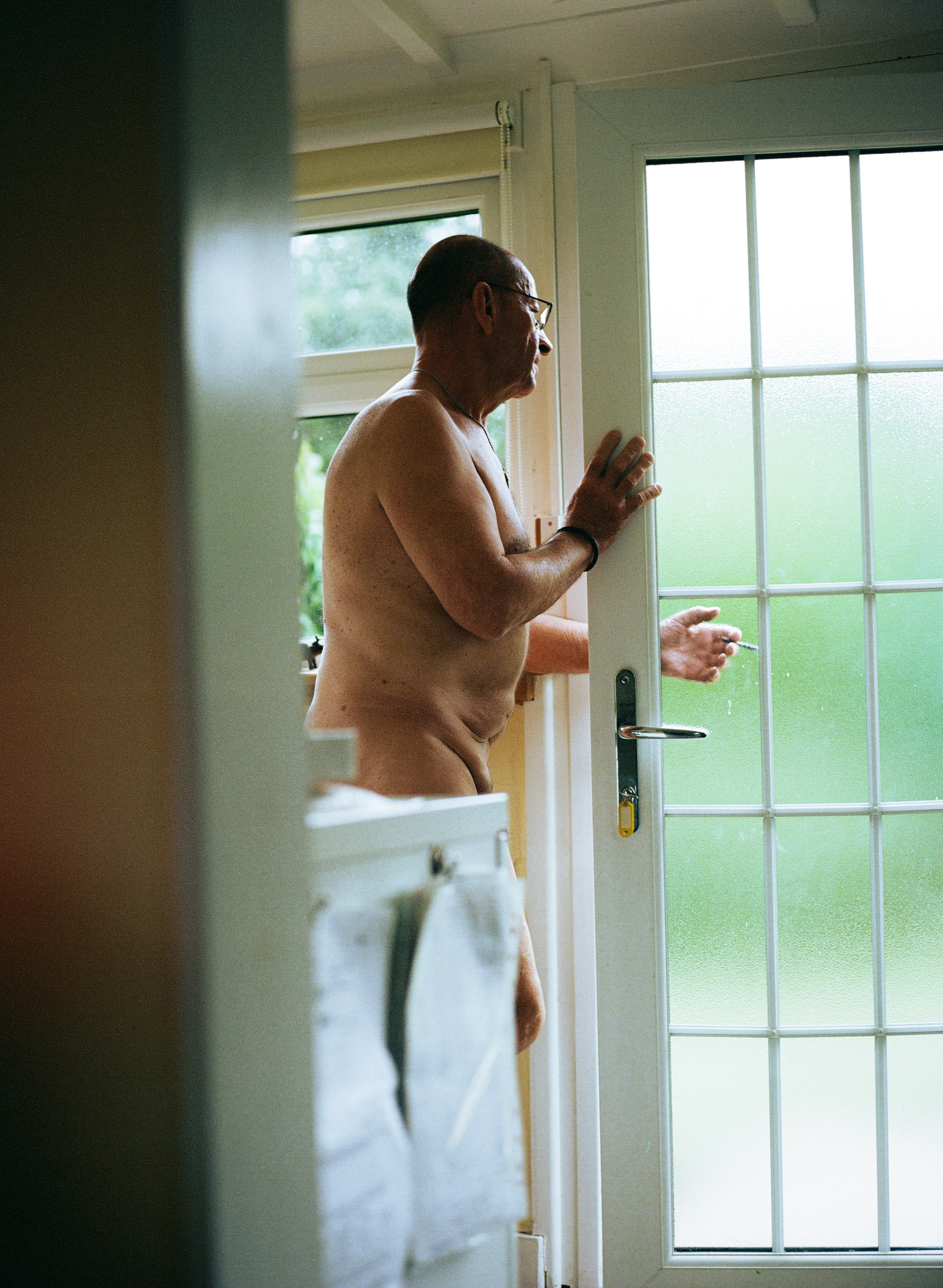 Man with glasses and white towel around waist standing at open glass door, viewed through doorway of bright room.