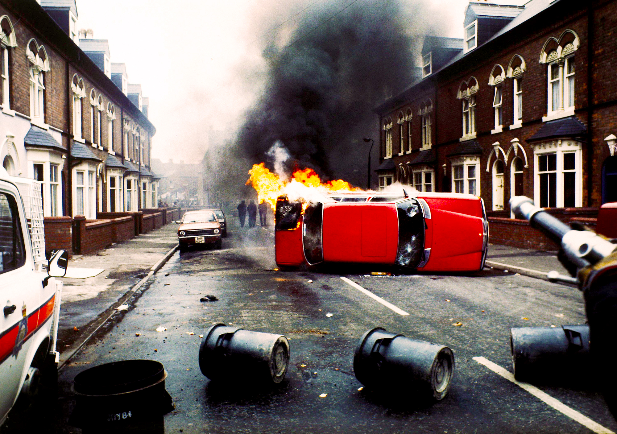 Overturned red van burning on residential street with black smoke, scattered tyres, and terraced houses either side.