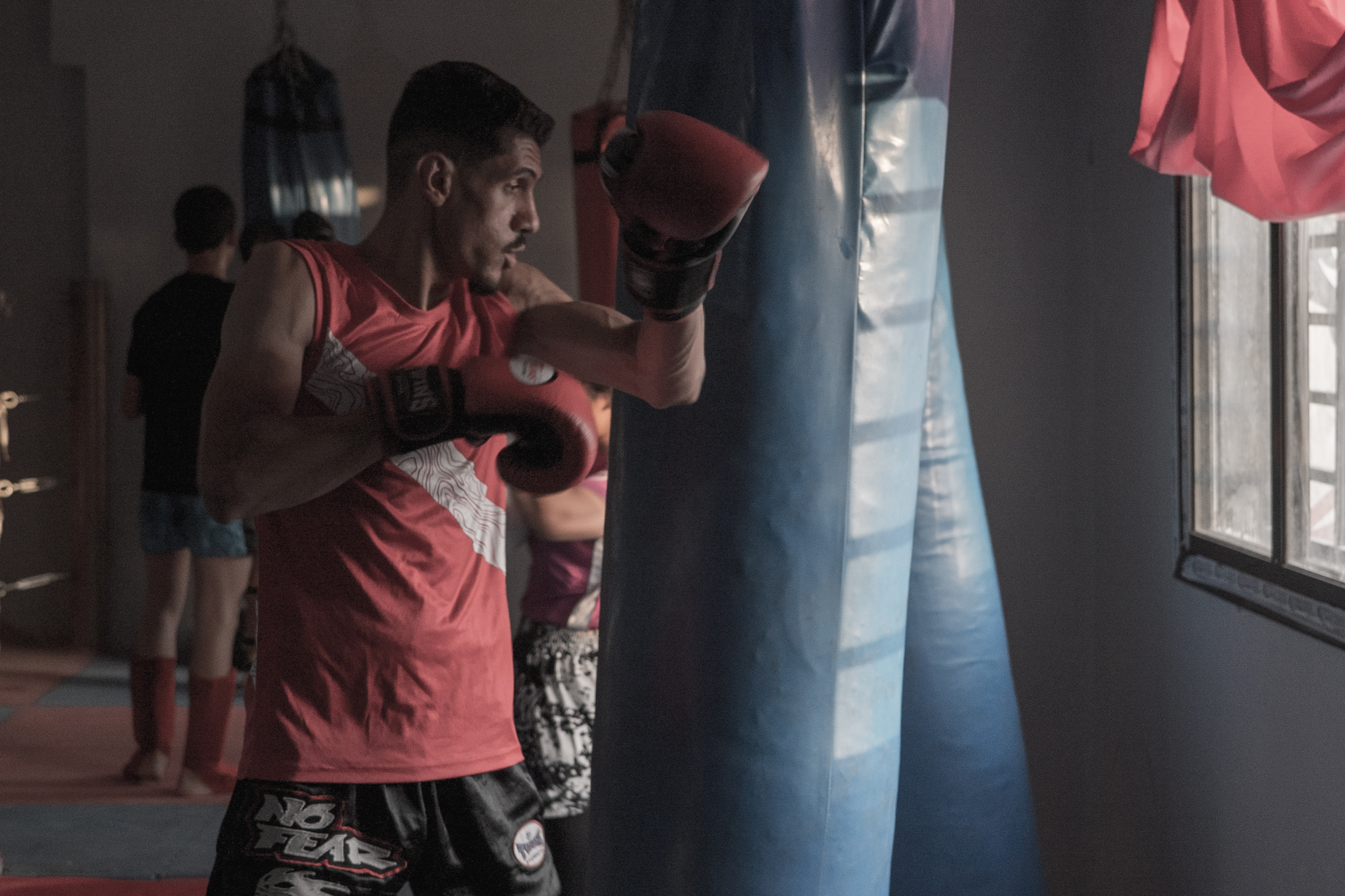 Woman in pink vest and black boxing gloves training with blue punching bag in gym with natural light from window.
