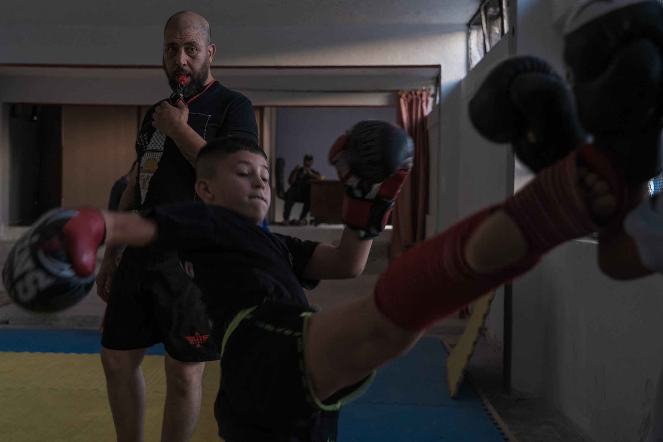 Man in black uniform restraining person in chair whilst others in red clothing assist in indoor setting with white walls.