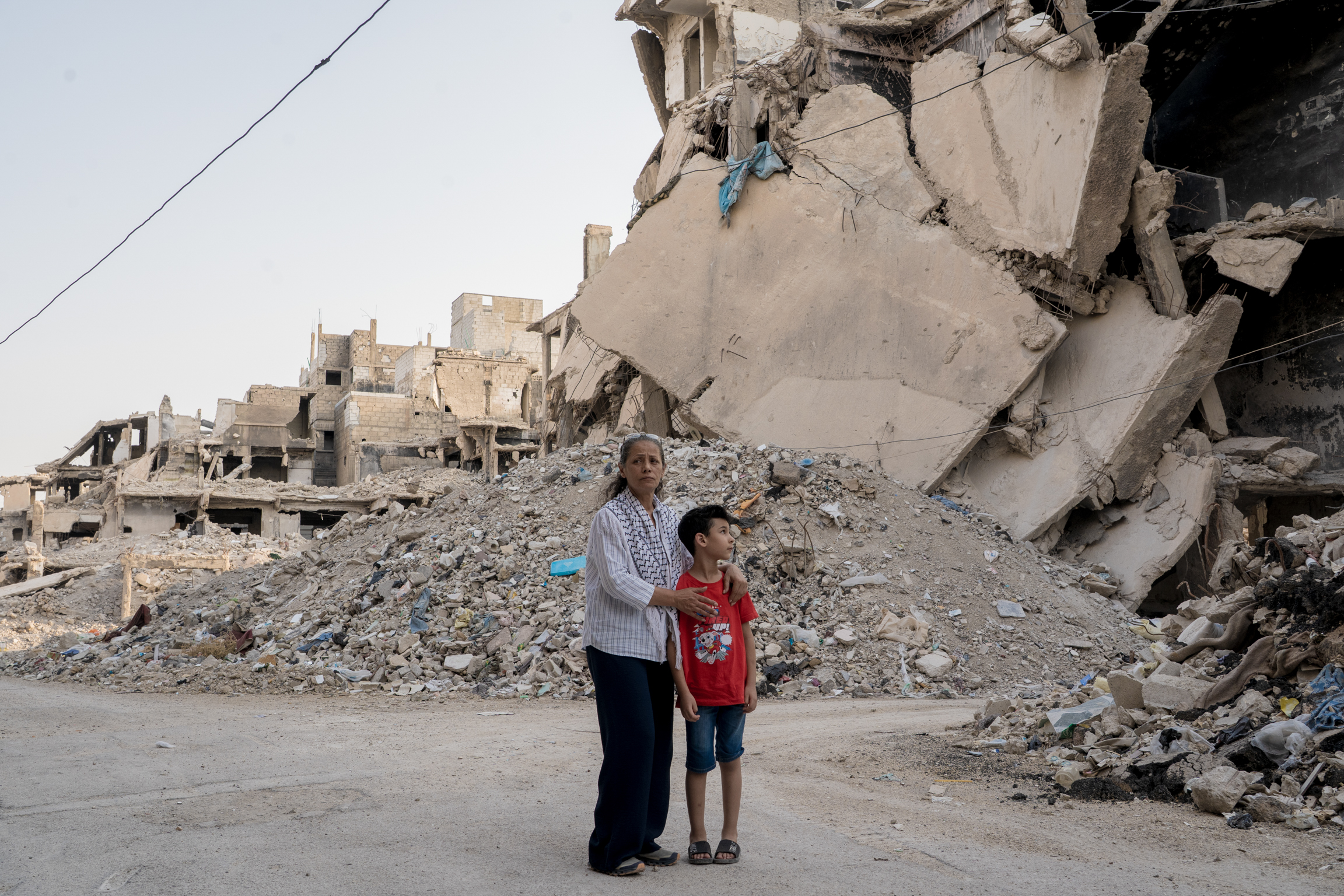 Elderly man and young boy standing amongst heavily damaged concrete buildings and rubble debris in urban area.