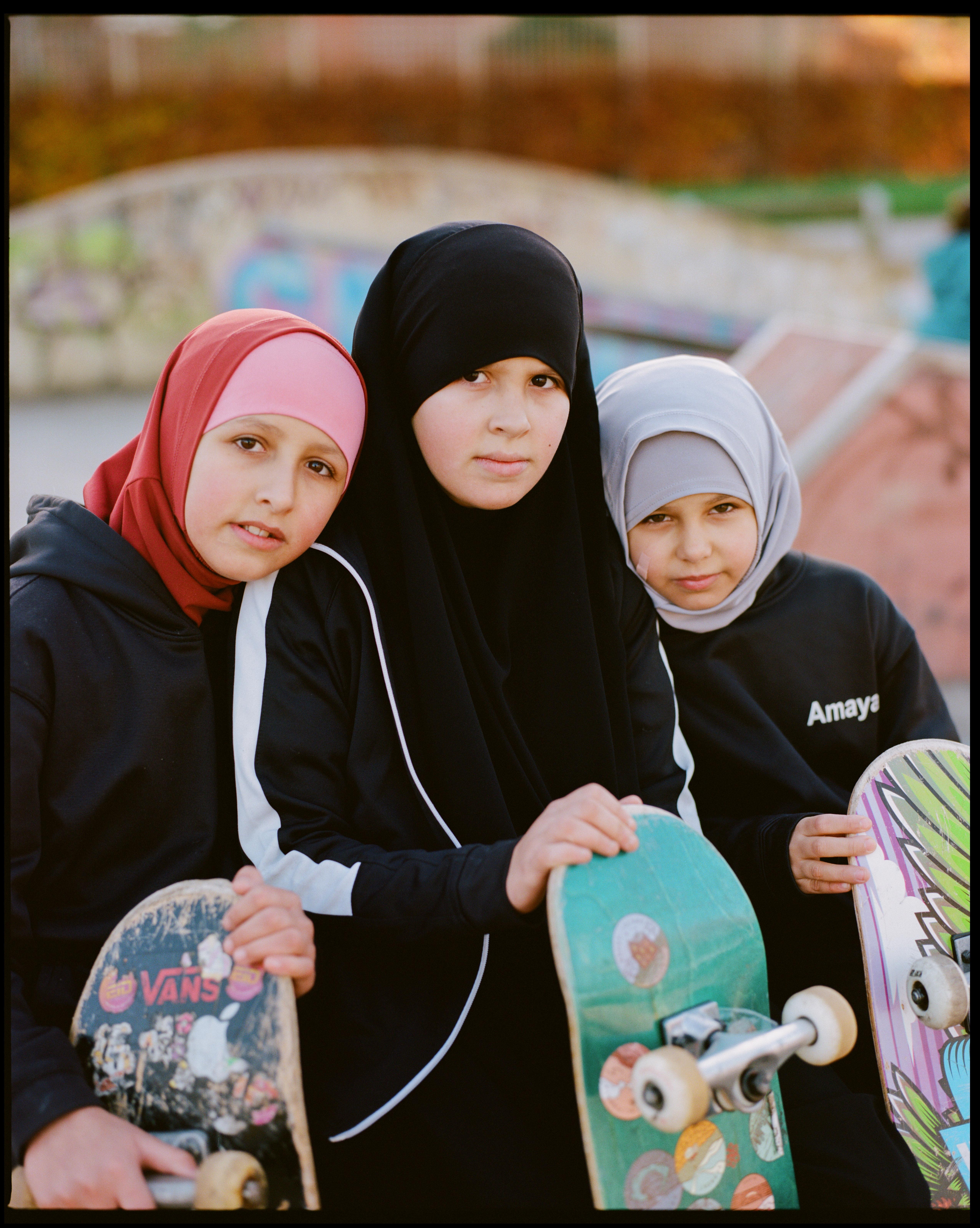 Three women in Islamic clothing, one wearing a pink headscarf, one wearing a black headscarf, and one wearing a grey headscarf, standing together and holding skateboards.