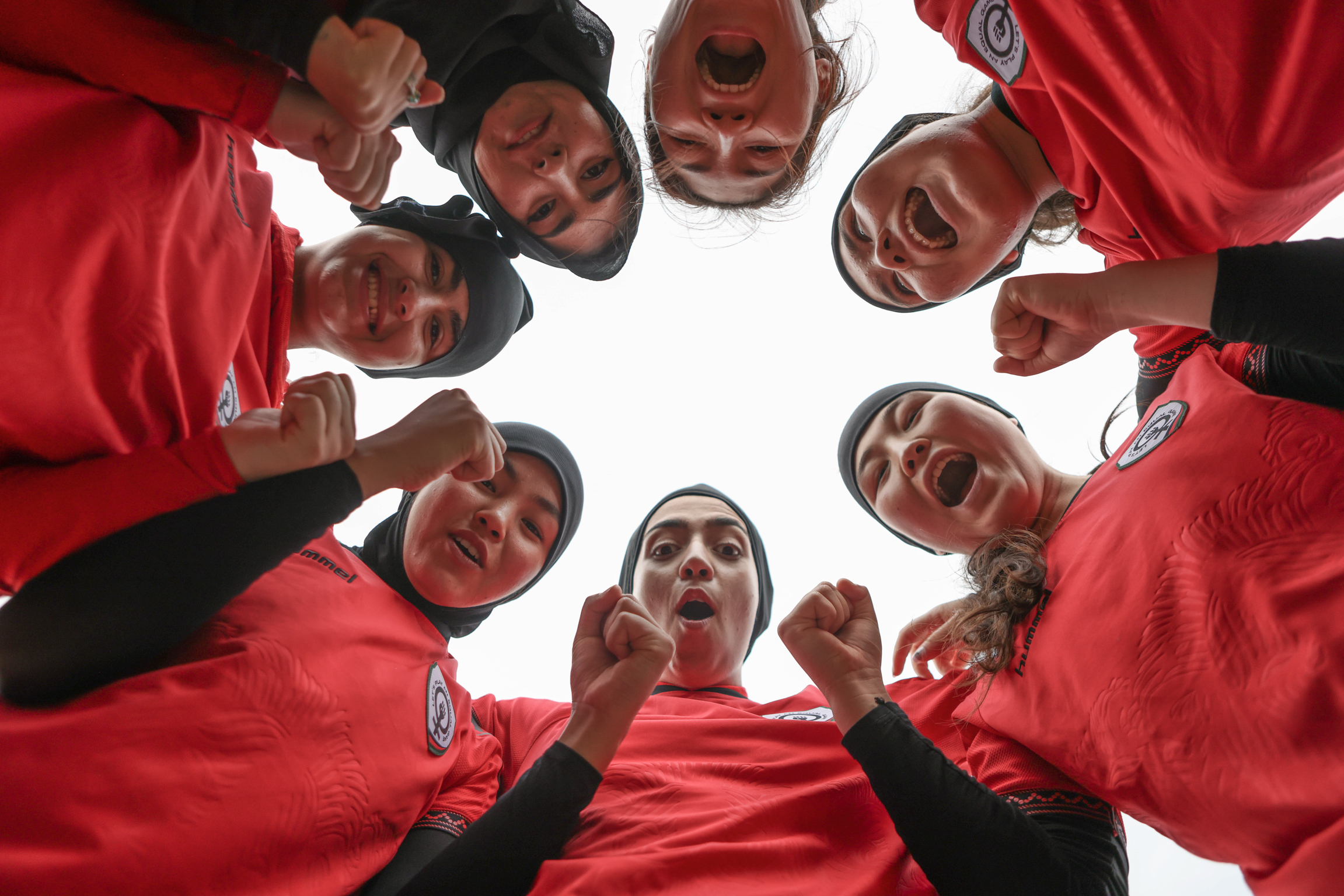 Eight people in red sports jerseys form a circle, heads together, looking down at camera from above against white background.