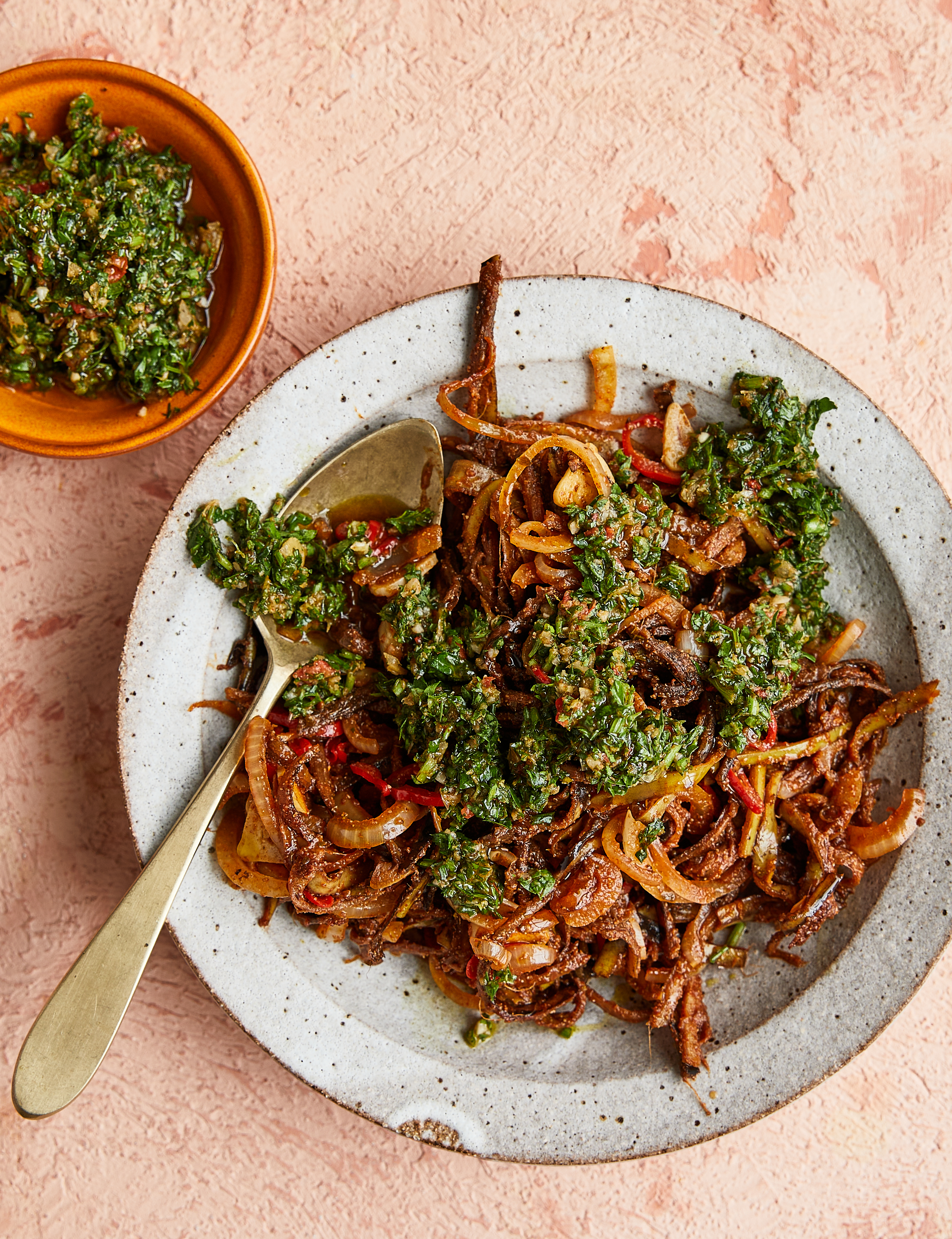 Speckled white plate with caramelised onions topped with green herb sauce, alongside terracotta bowl of herbs on pink surface.