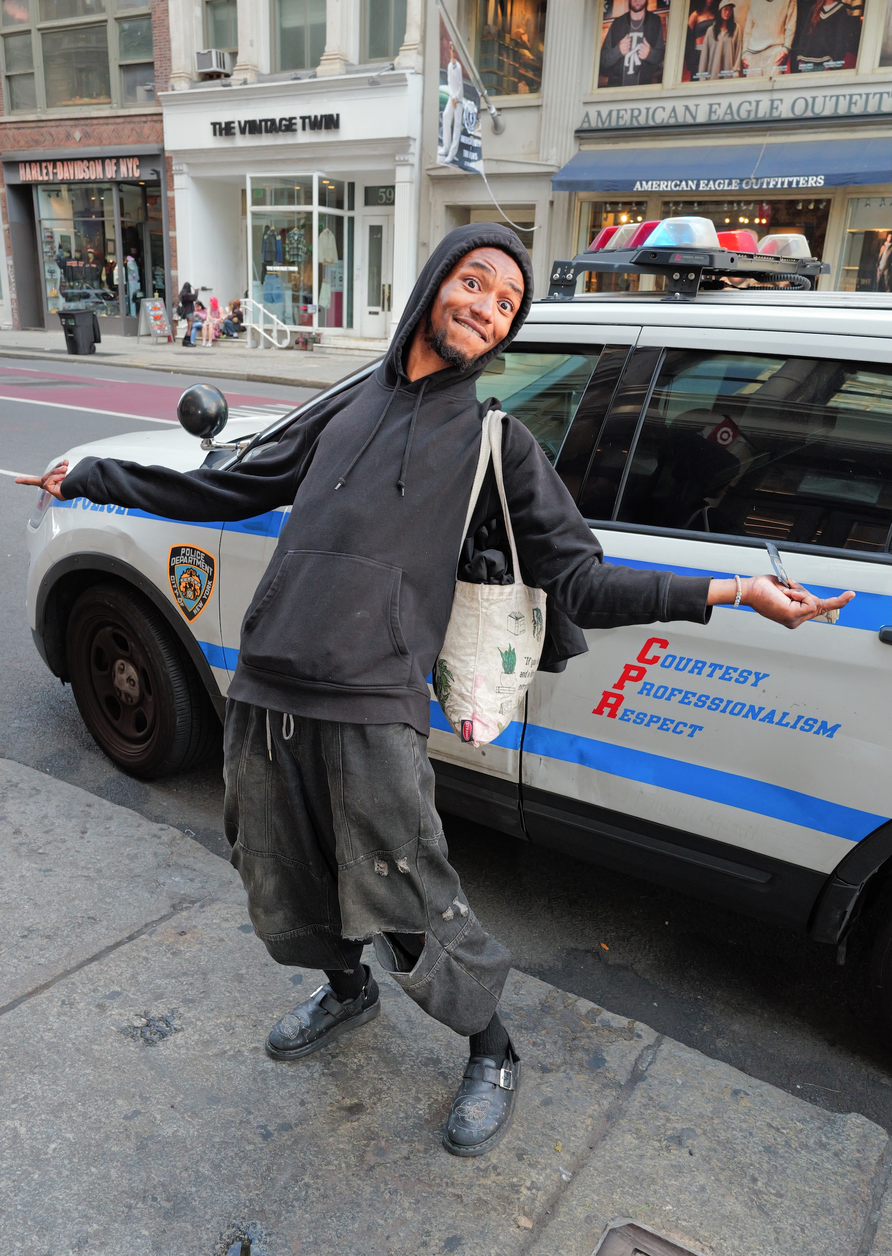 Person in black hoodie and grey trousers standing beside white police car with blue stripes on city street with shops in background.