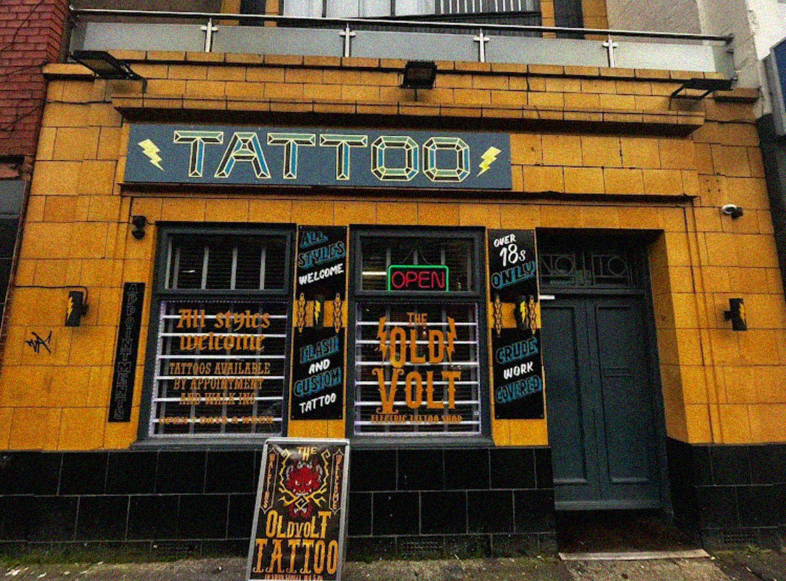 Yellow brick tattoo shop with black signage, large windows displaying artwork, and neon "OPEN" sign in storefront.