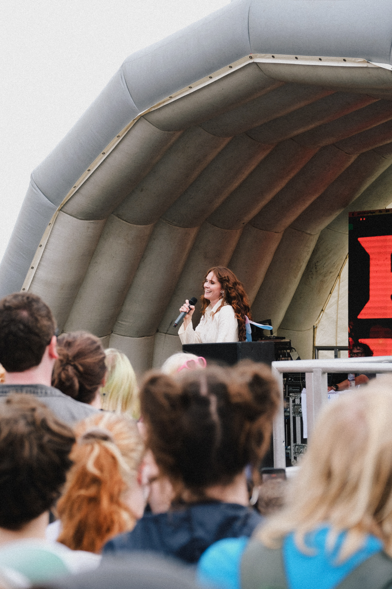 Woman in white shirt performing on stage under grey inflatable canopy with audience watching from below.
