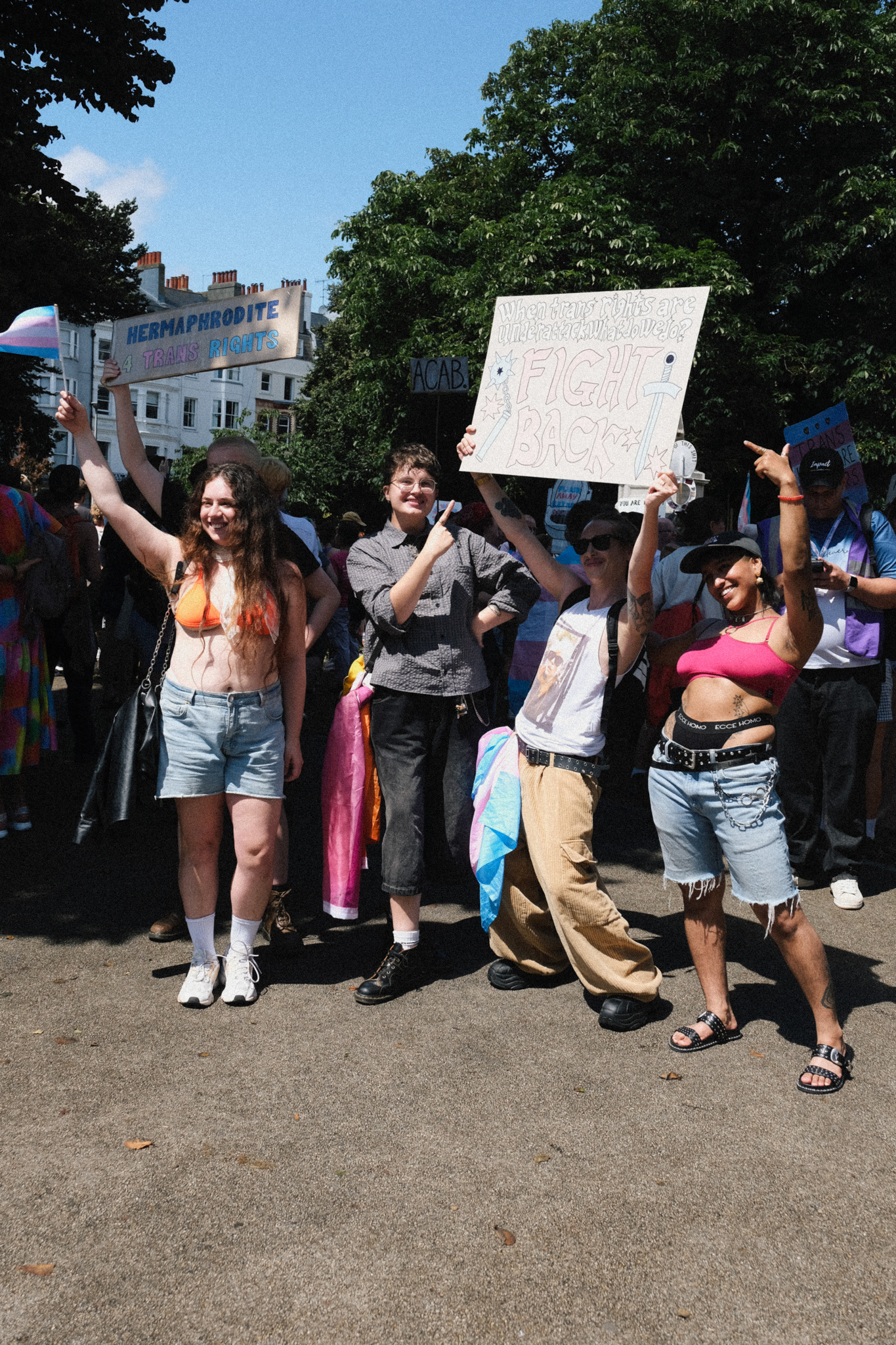 Group of protesters holding signs and flags on sunny street, surrounded by green trees with white buildings in background.