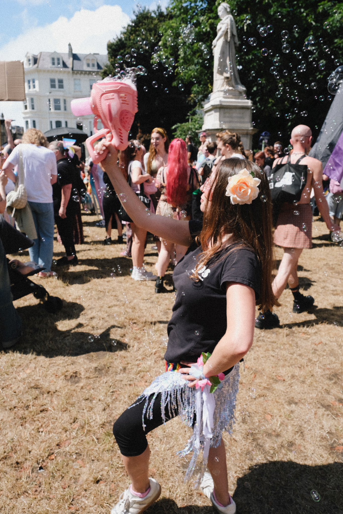 Woman in black top and leggings with tinsel skirt holds pink balloon at outdoor festival. Crowd and statue visible in background.