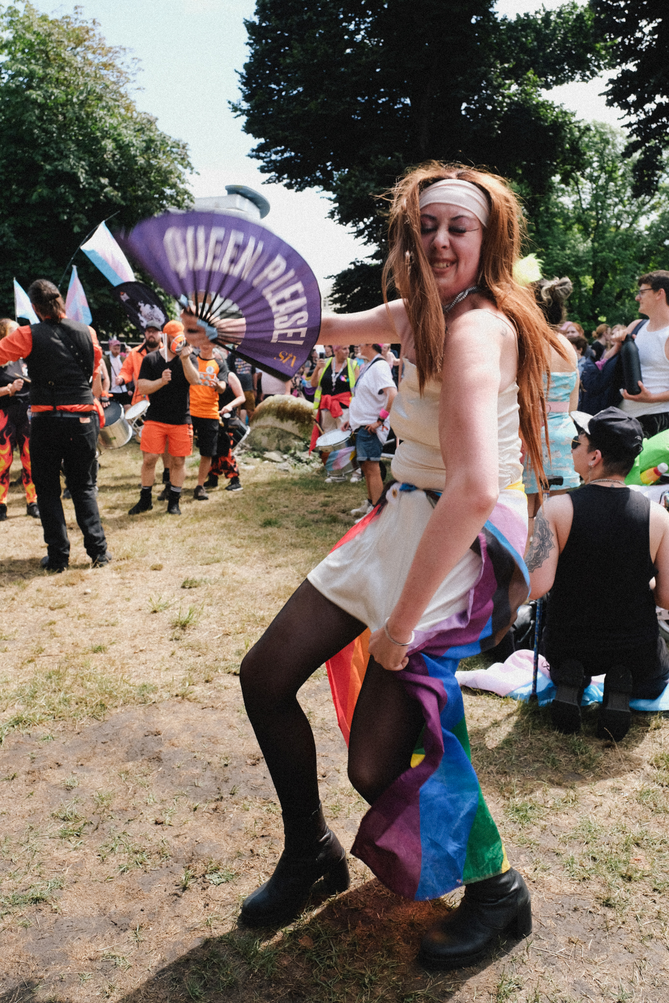 Person with long blonde hair holding purple fan reading "QUEEN BEE" at outdoor Pride event, wearing rainbow cape and headband.