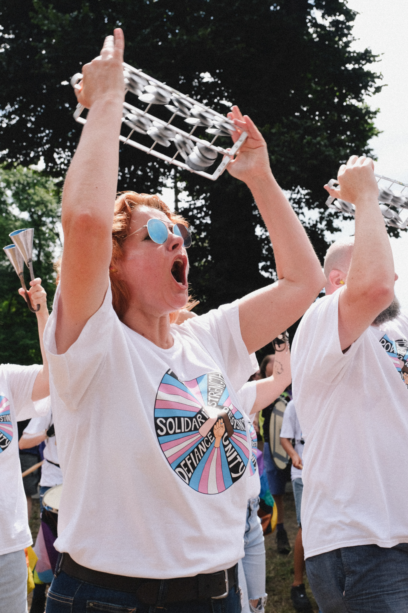 Woman in white t-shirt and sunglasses holds trophy aloft with both hands, mouth open in celebration, surrounded by cheering people.