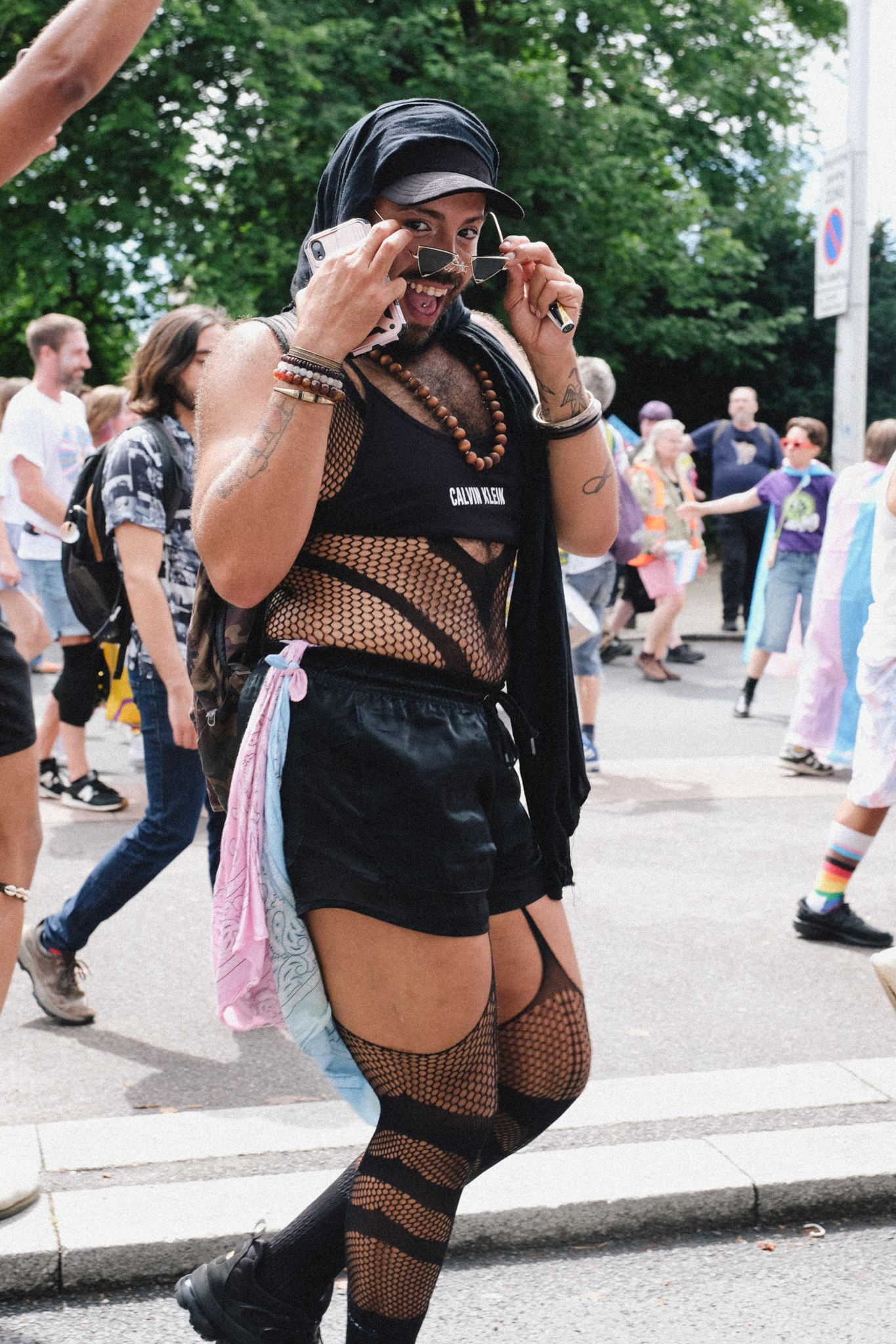 Person in black leather cap and fishnet outfit taking photos with small camera at outdoor street event with crowd in background.