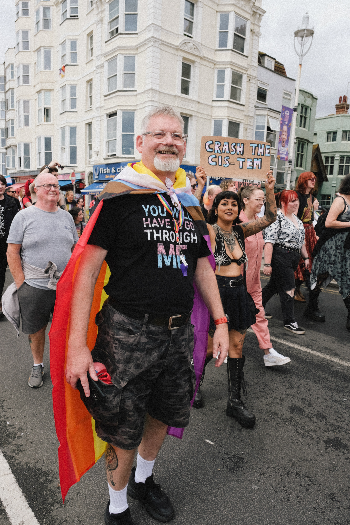 Man wearing rainbow flag cape and black t-shirt walks with crowd on street lined with white Victorian buildings during Pride parade.