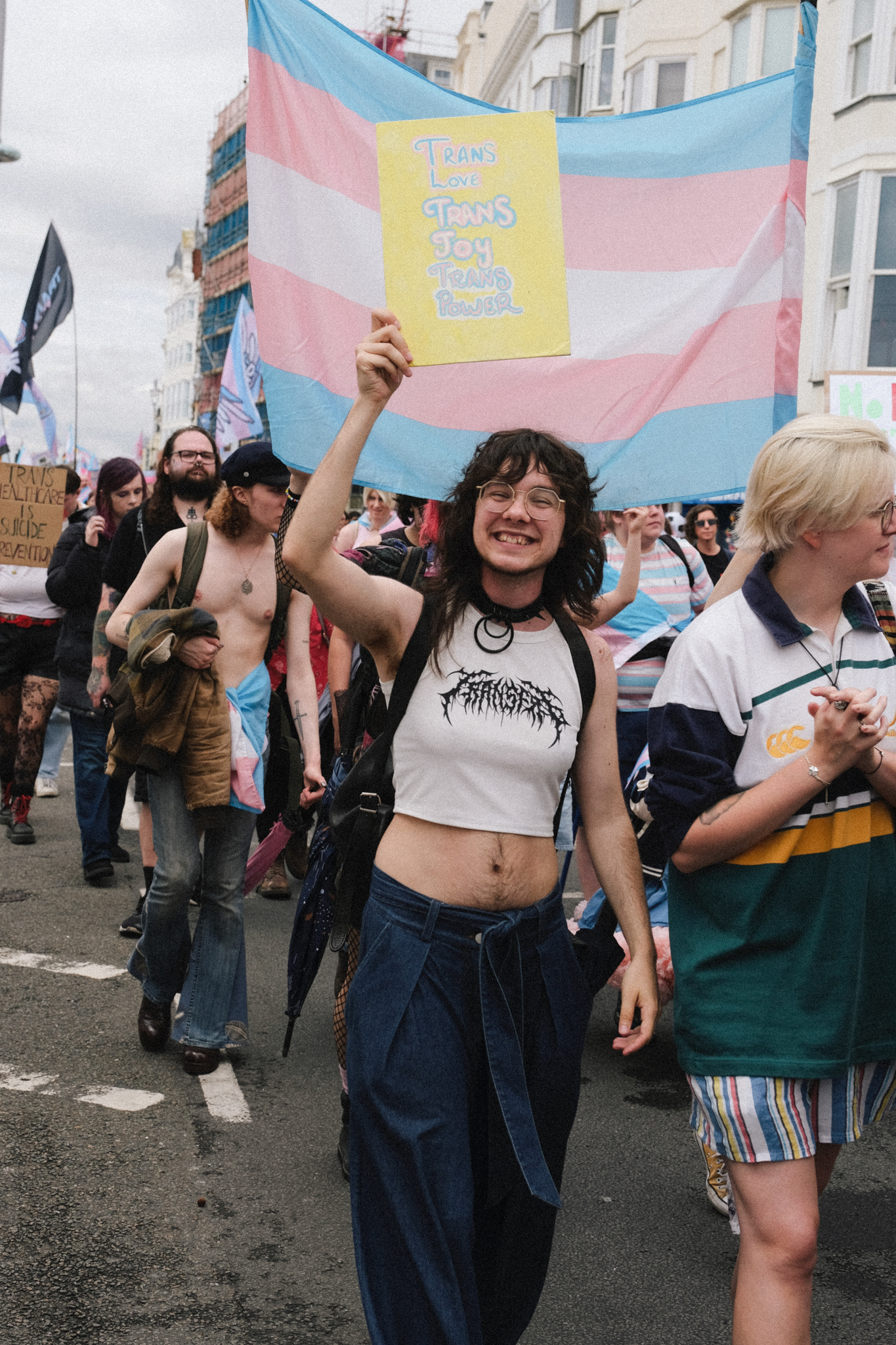 Person holding yellow sign at protest march with large transgender flag in background, crowd on street with white buildings.