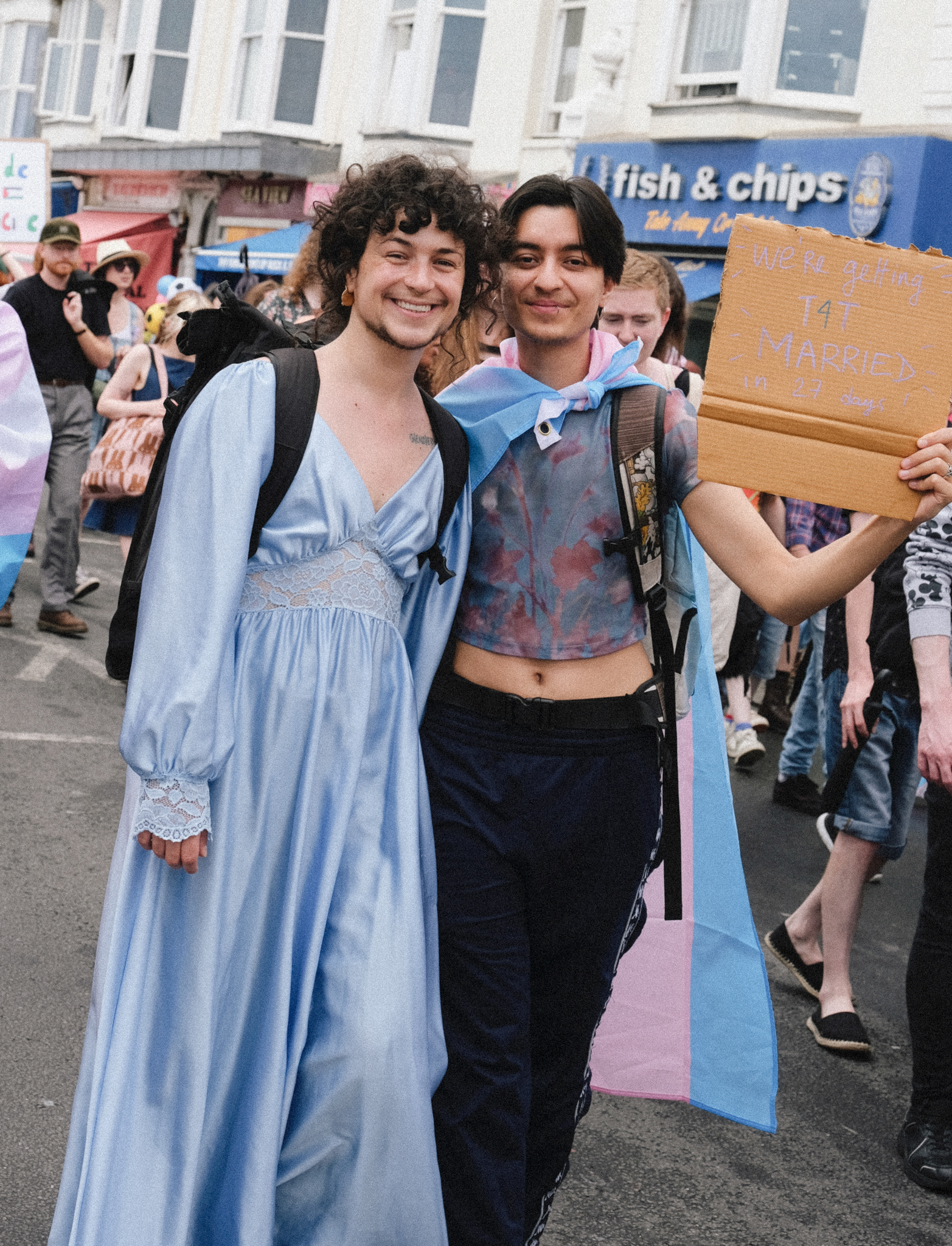 Two people at street demonstration, one in blue period dress with curly wig, other in tie-dye top. Protesters and fish & chips shop behind.