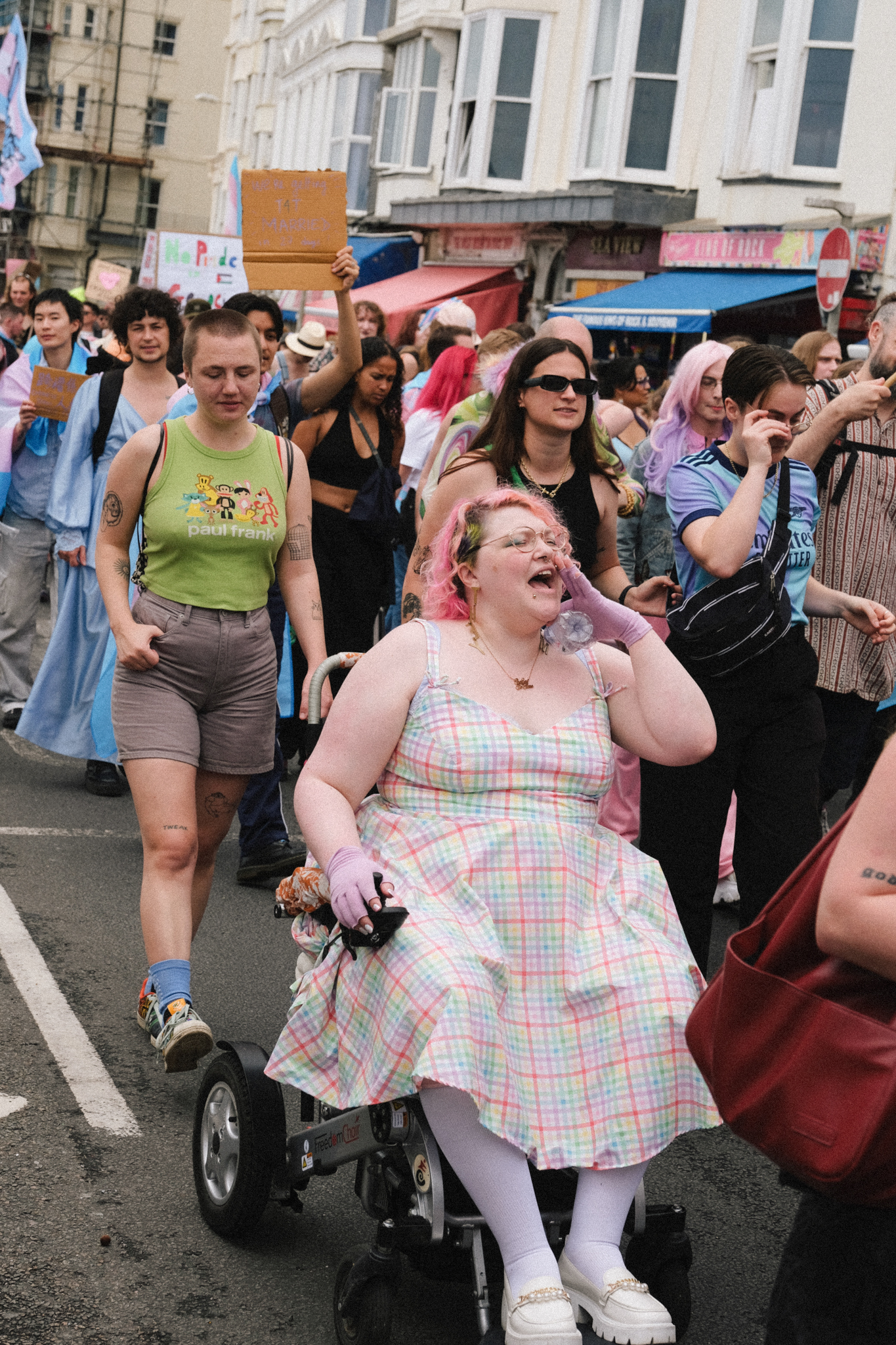 Woman in wheelchair wearing pastel plaid dress at crowded street demonstration with protesters holding signs and colourful banners.