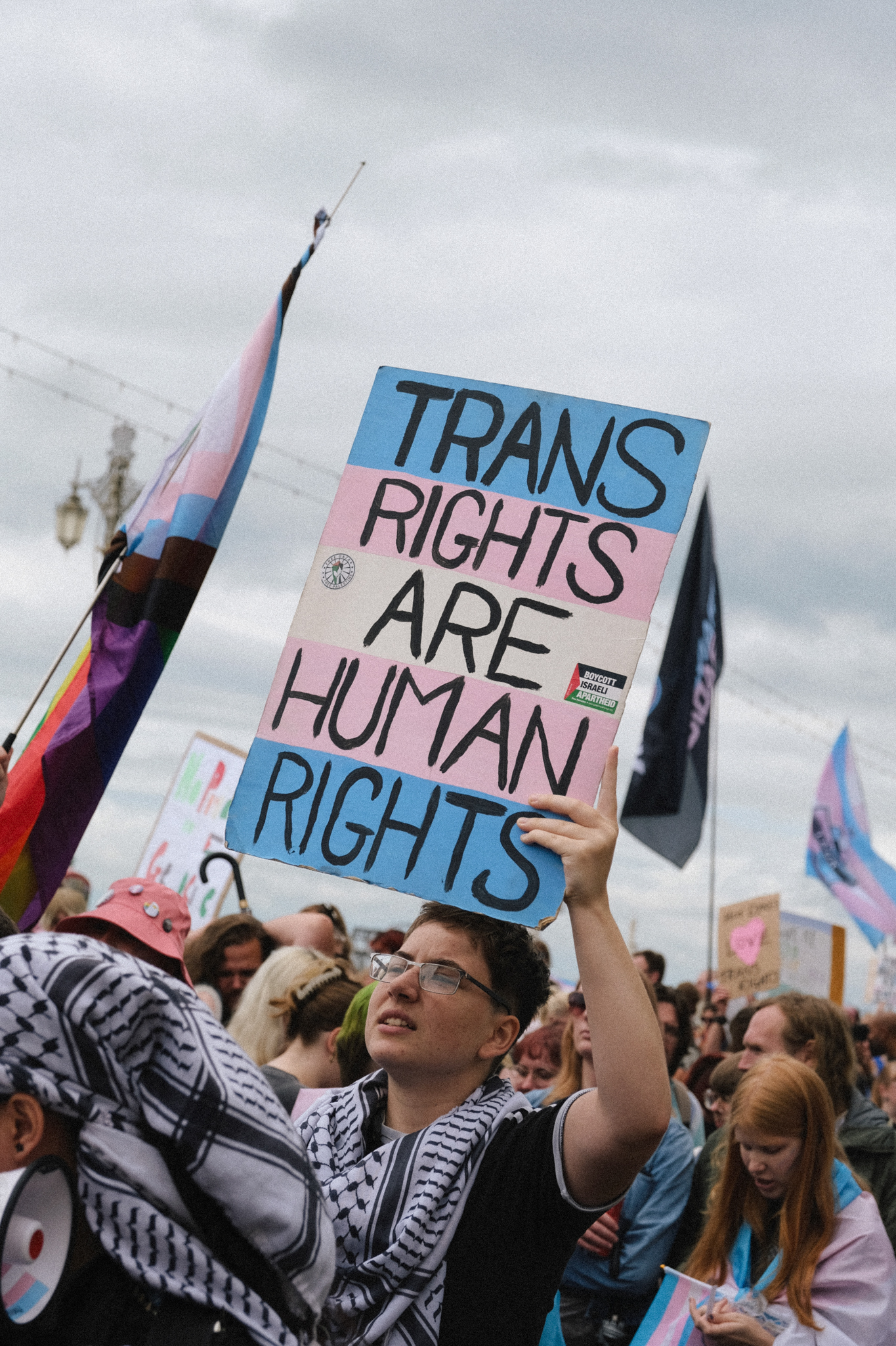 Person holding white placard with blue and black text "TRANS RIGHTS ARE HUMAN RIGHTS" at crowded outdoor demonstration with flags.