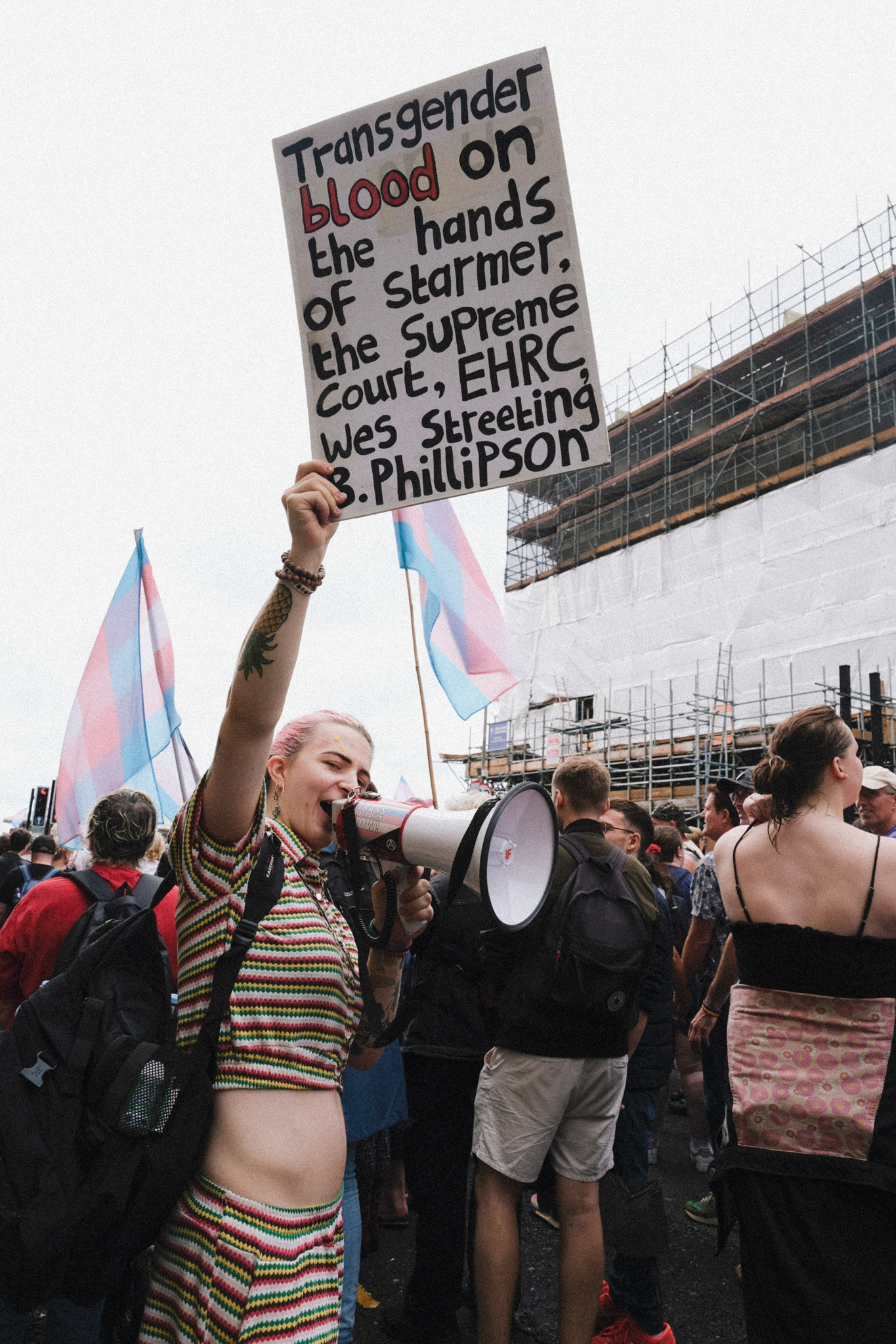 Protest crowd with person holding white placard with black text, blue and pink transgender flags visible, construction scaffolding in background.
