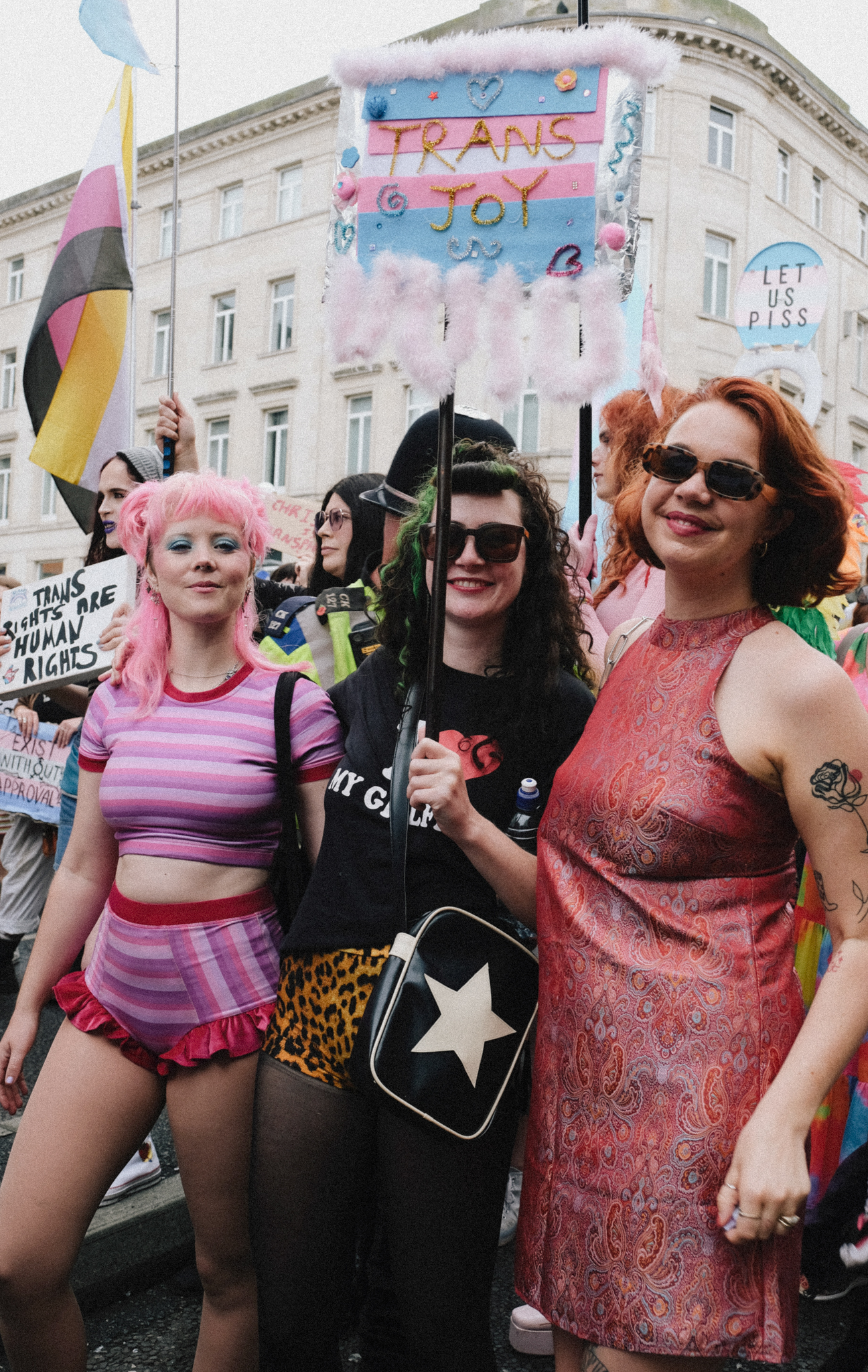 Three people at pride march holding colourful signs including "TRANS & JOY" banner. Pink-haired person in striped outfit, person in black, person in red dress.
