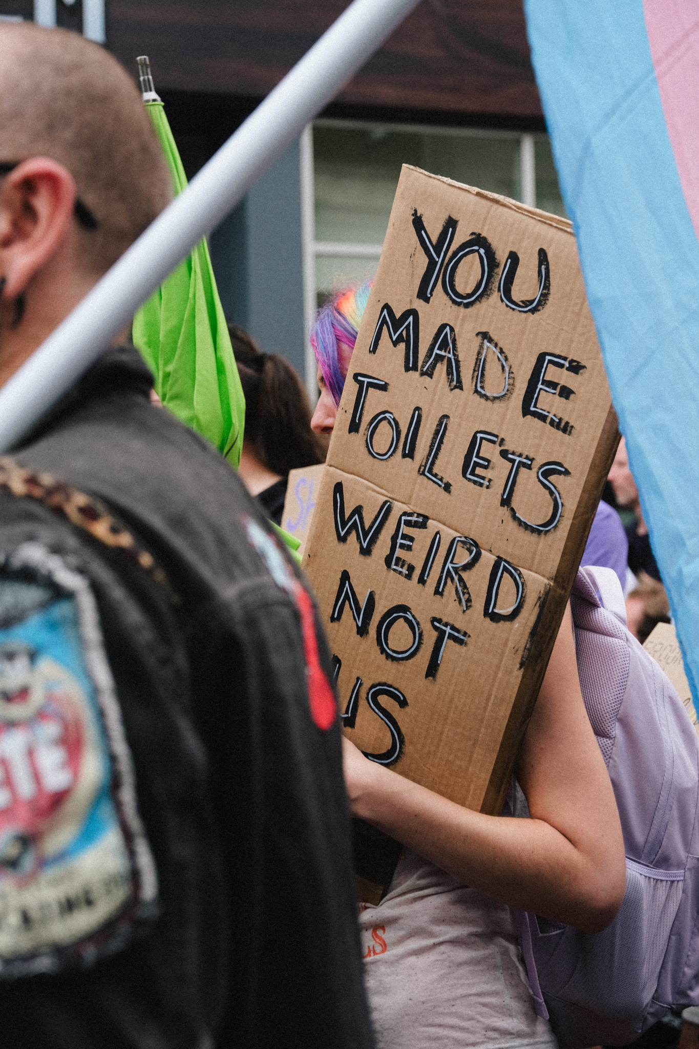 Person holding cardboard sign reading "YOU MADE TOILETS WEIRD NOT US" at protest with colourful flags and other demonstrators.