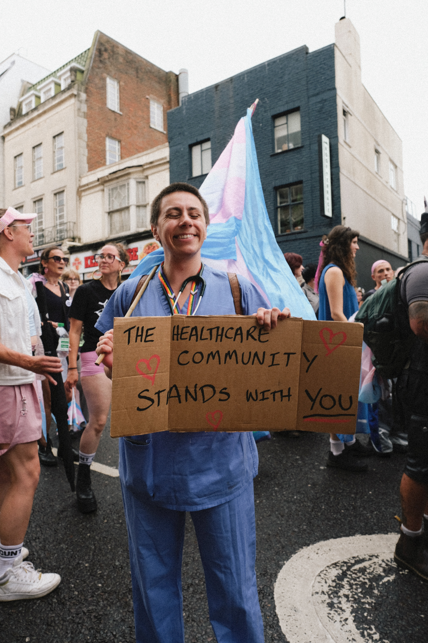Person in blue medical scrubs holds cardboard sign reading "The Healthcare Community Stands With You" at street demonstration with transgender pride flag and crowd behind.