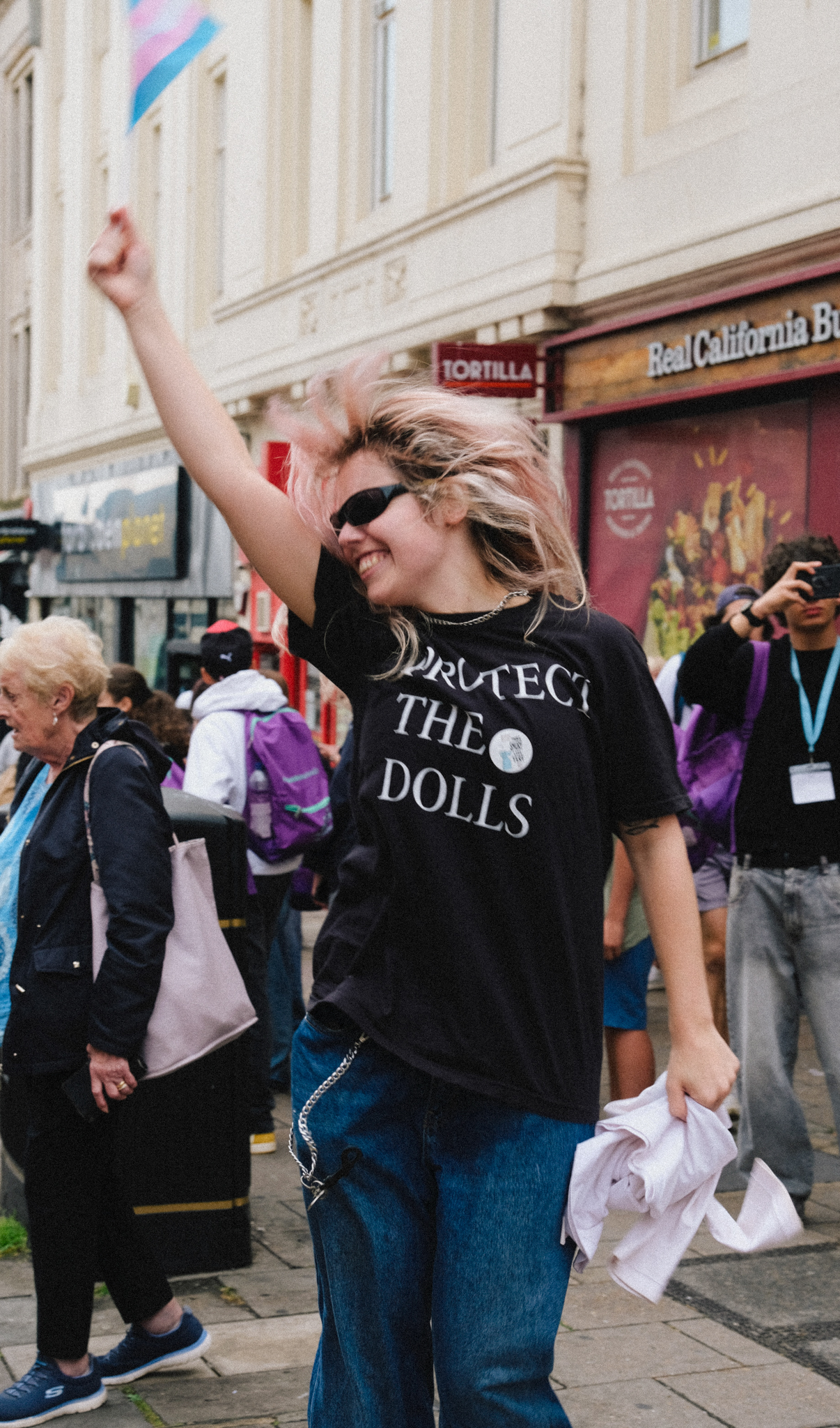 Woman in sunglasses and "Protect the Dolls" t-shirt raises arm at street demonstration, crowd and shops visible behind.