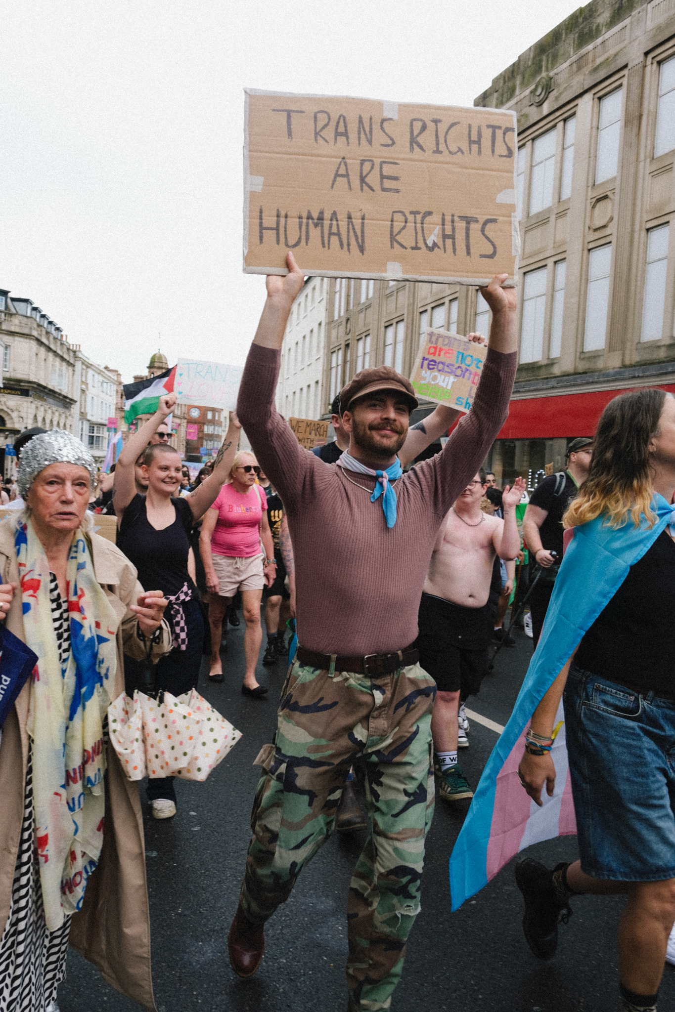 Person in camouflage trousers holding cardboard sign reading "TRANS RIGHTS ARE HUMAN RIGHTS" at street demonstration with crowd and buildings.