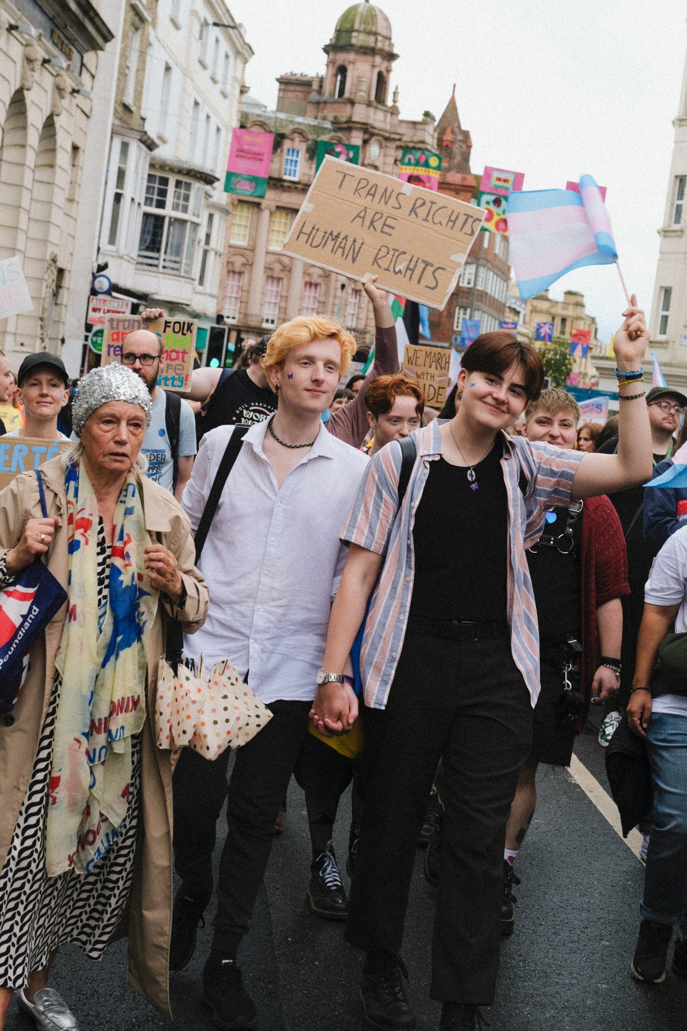 Crowd of protesters holding signs including "Trans Rights are Human Rights" walking down street with colourful buildings and clock tower.