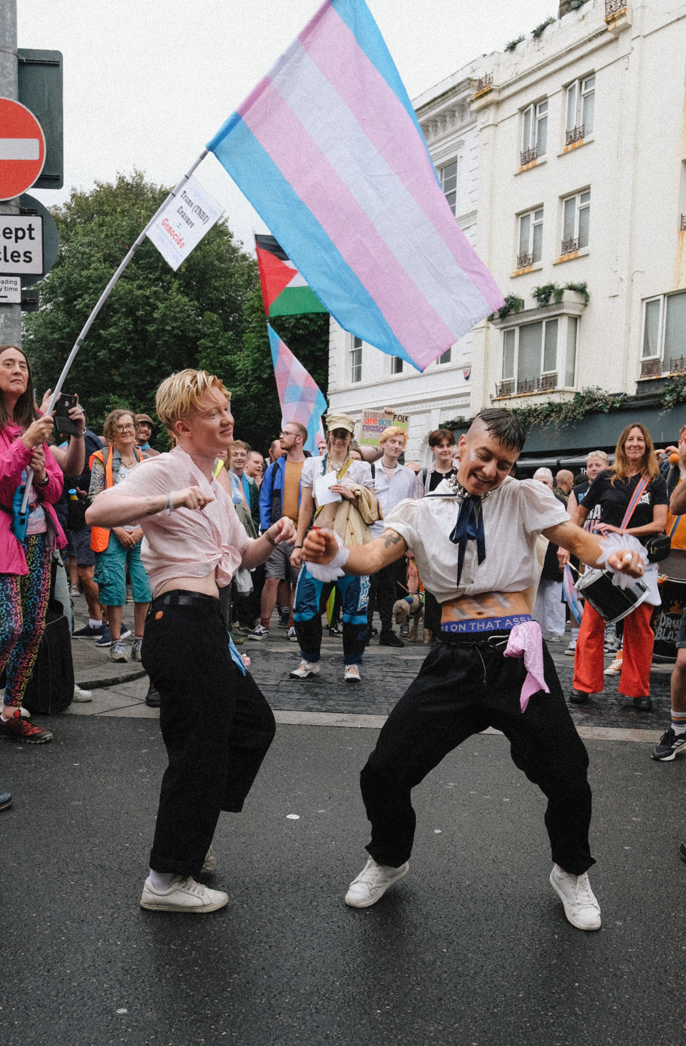 Two people dancing in street holding transgender pride flag with light blue, pink and white stripes. Crowd watching behind them.