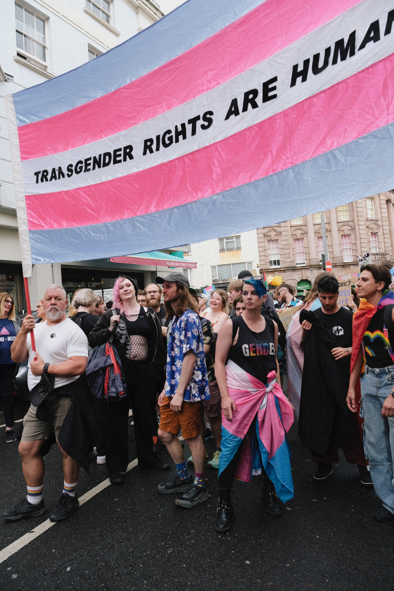 Large banner with pink and blue stripes reading "TRANSGENDER RIGHTS ARE HUMAN" held above crowd of protesters on street.