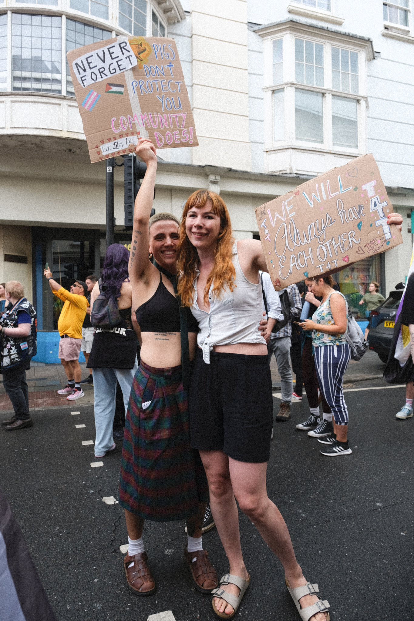 Two women holding handwritten cardboard protest signs on a street with white buildings and crowd in background.