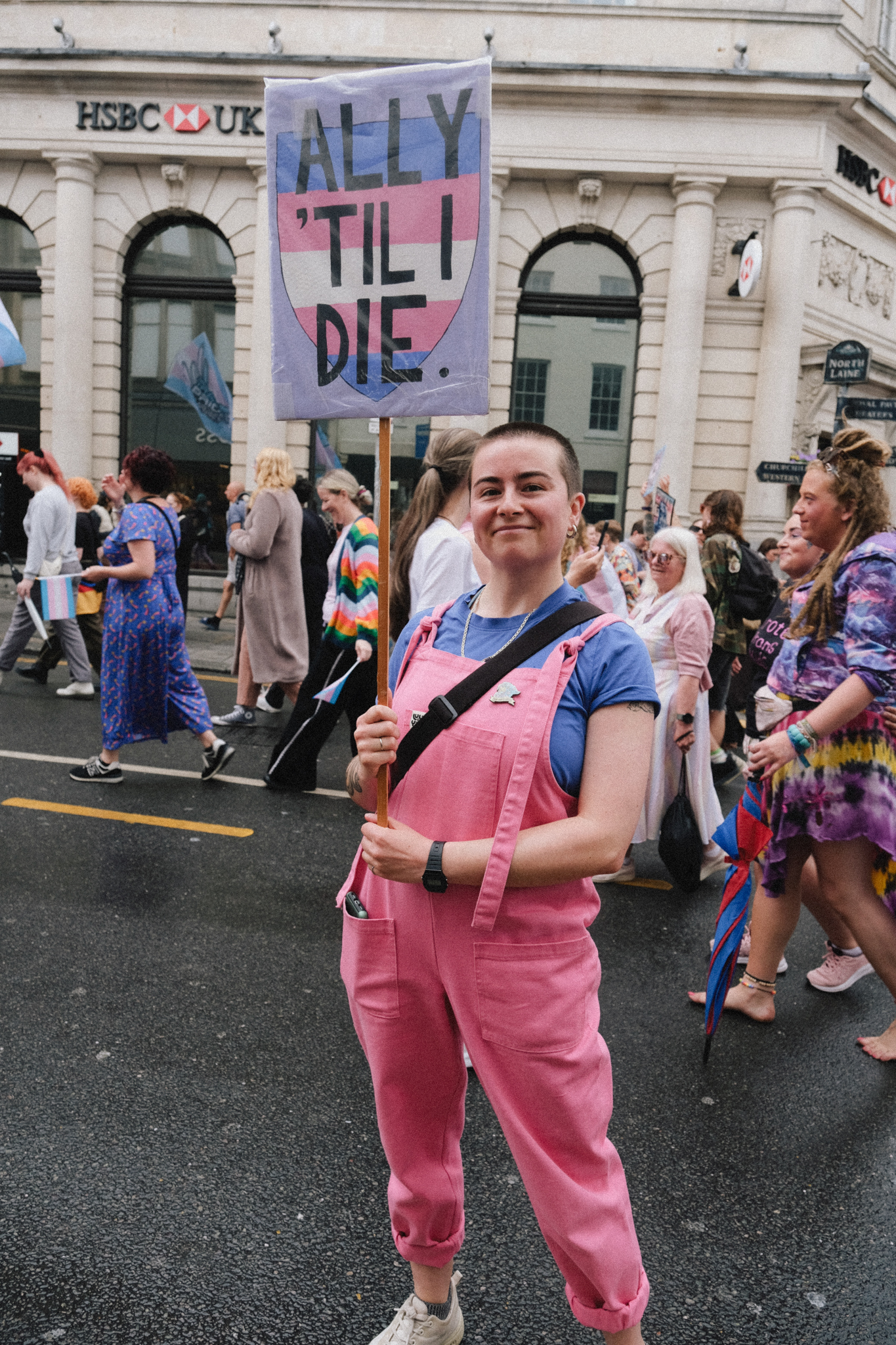 Woman in pink dungarees holding protest sign reading "ALLY 'TIL I DIE" at street demonstration outside HSBC bank building.