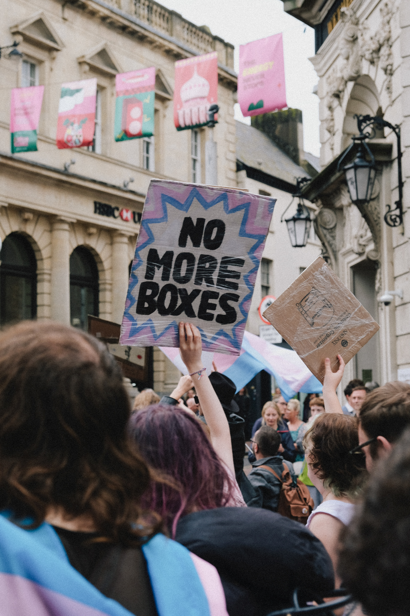Protesters holding signs including "NO MORE BOXES" in front of historic stone buildings with classical columns and arched windows.