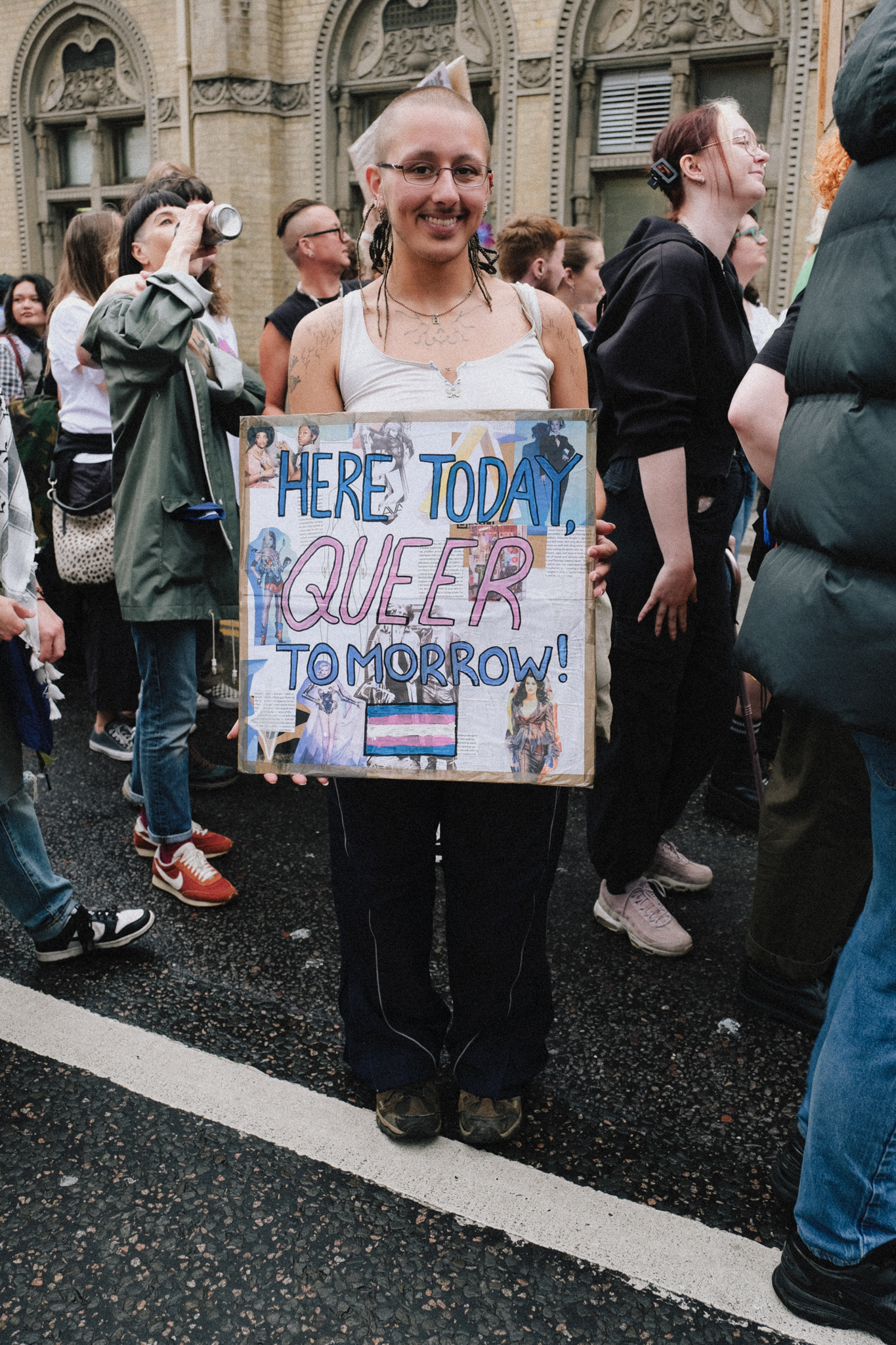 Person at Pride event holding handmade sign reading "HERE TODAY, QUEER TOMORROW!" with transgender flag, surrounded by crowd.