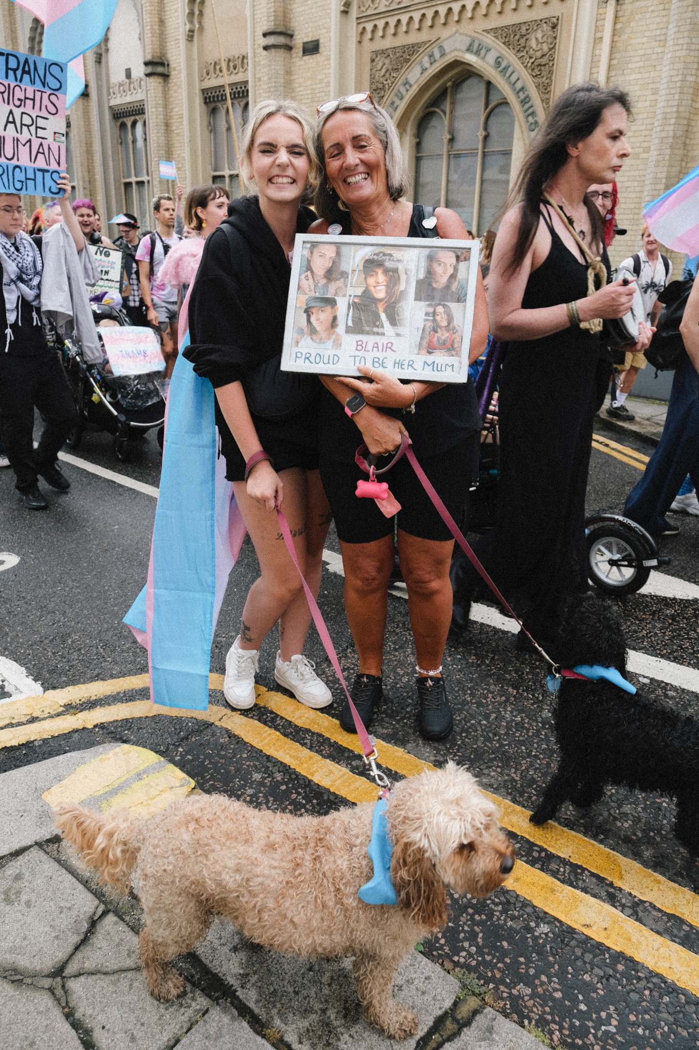 Two women at trans rights protest holding sign with photos, one with walking stick, small dog on lead, crowd with flags behind.