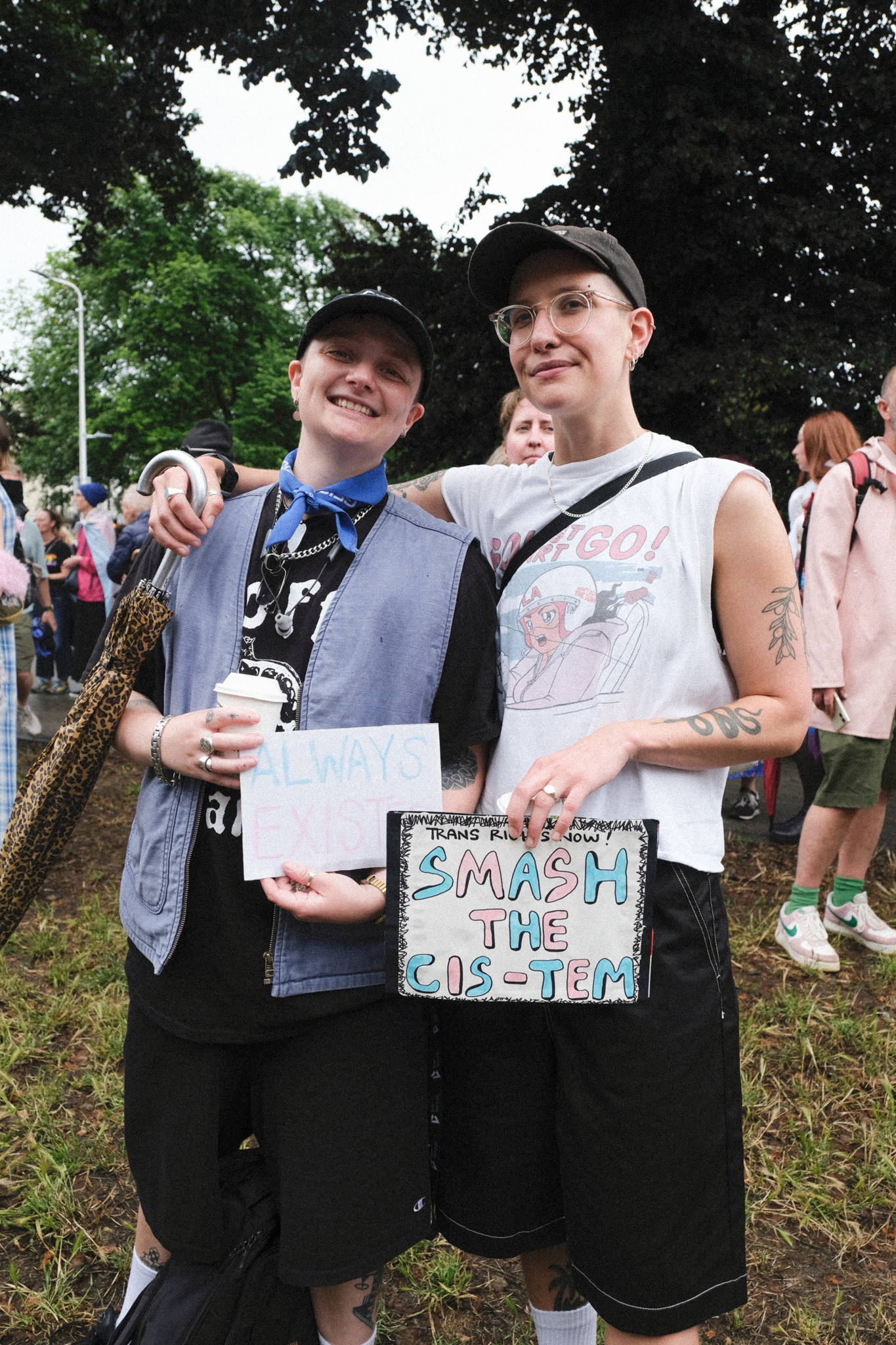 Two people with arms around each other holding a handmade sign reading "SMASH THE CIS-TEM" at an outdoor gathering with trees and crowd.