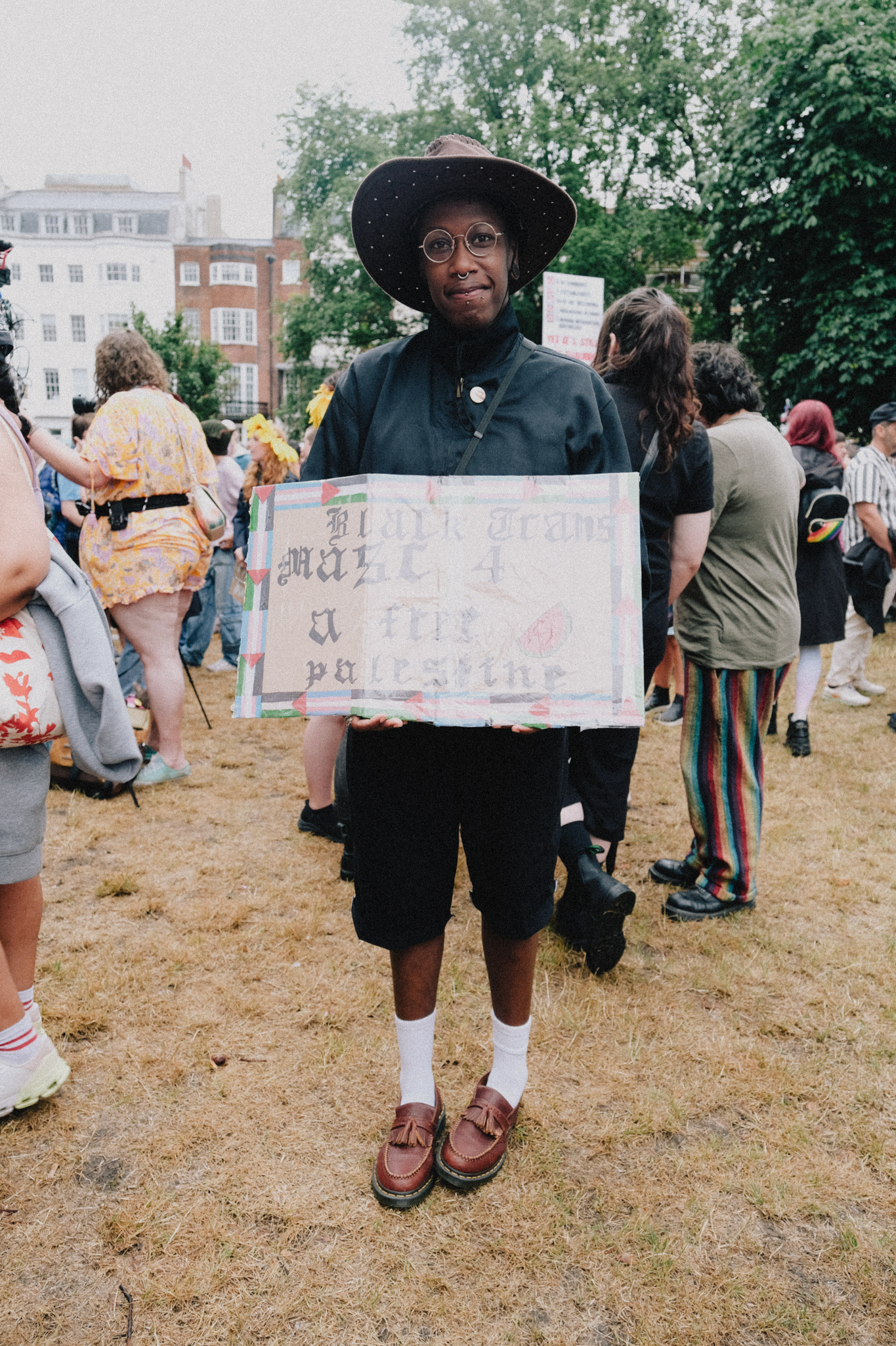 Person in black hat and clothing holds protest sign at outdoor gathering, with crowd and Georgian buildings in background.