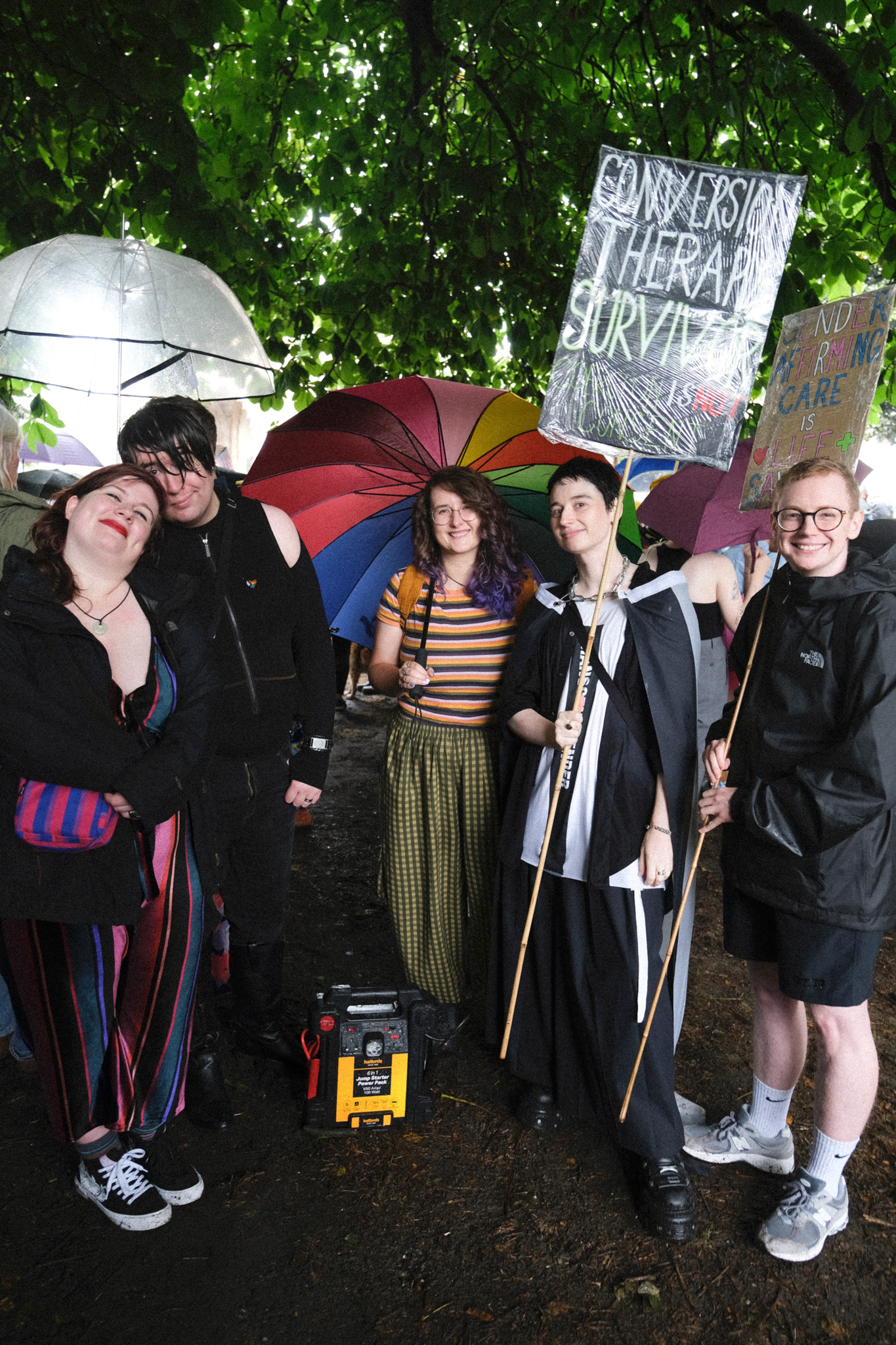 Group of people outdoors with umbrellas and protest signs including "conversion therapy survivor" under green trees.