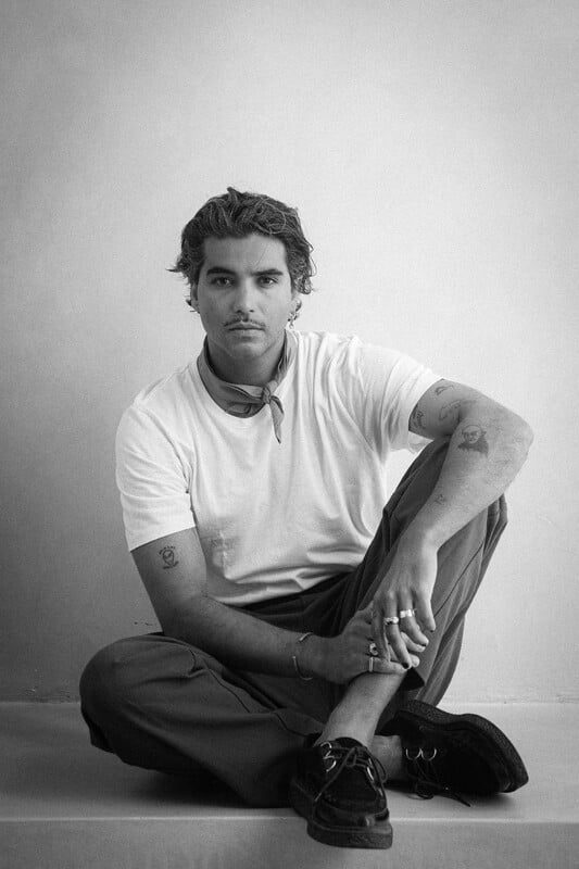 Black and white portrait of young man with curly hair sitting cross-legged, wearing white t-shirt and dark trousers with trainers.