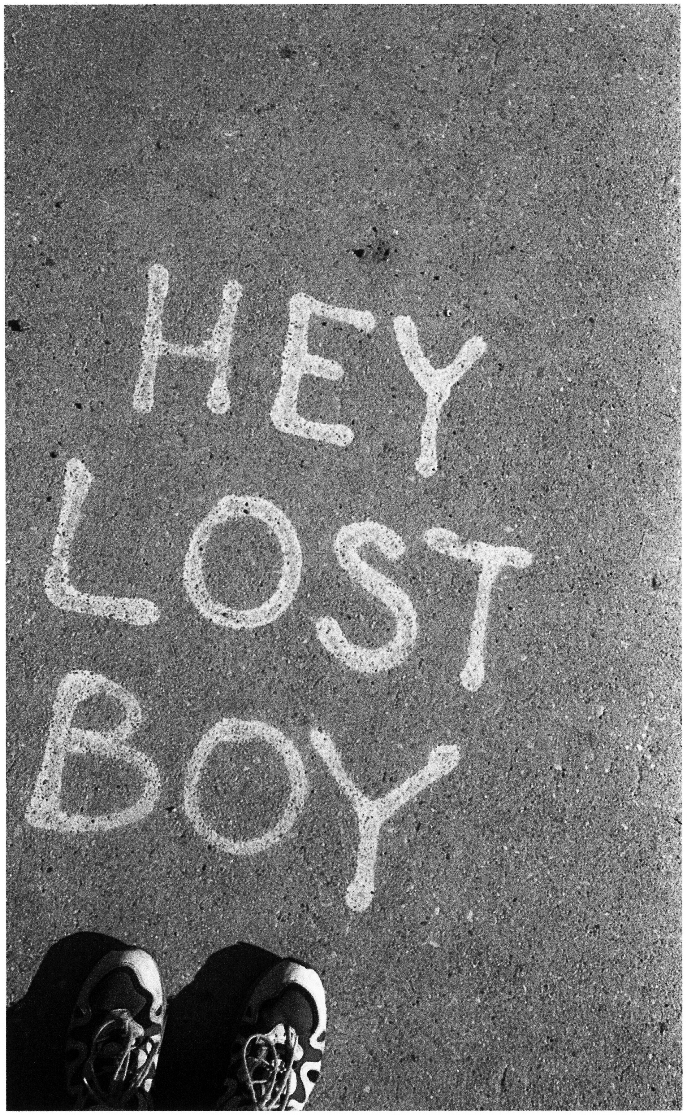 White chalk text "HEY LOST BOY" on grey pavement with black shoes visible at bottom edge.