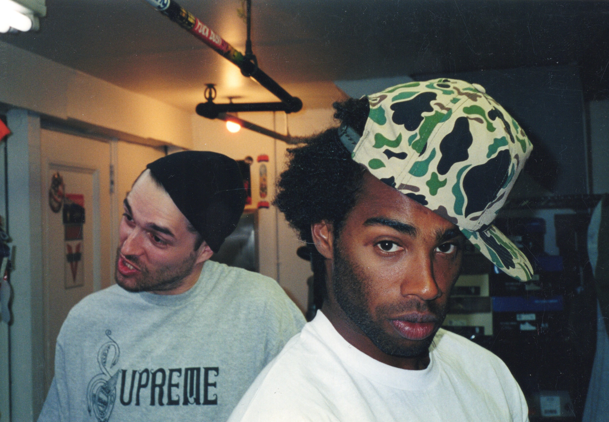 Two men in white tops, one wearing black beanie, other in green camouflage cap, standing in dimly lit indoor space.