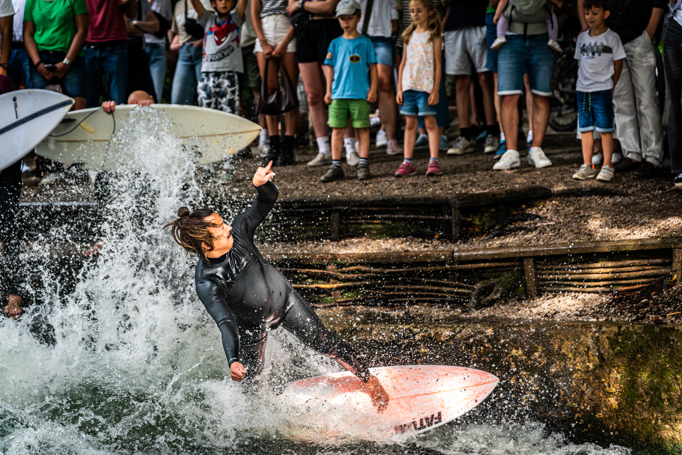 Surfer in black wetsuit on pink surfboard creating splash in shallow water with crowd of children watching from stone steps above.