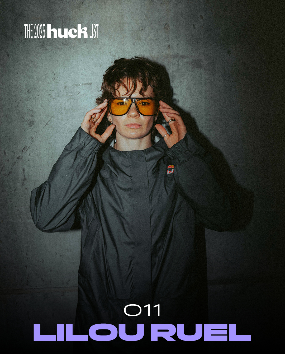 Young man with curly hair wearing orange sunglasses and dark jacket, hands positioned near his face against grey textured background.