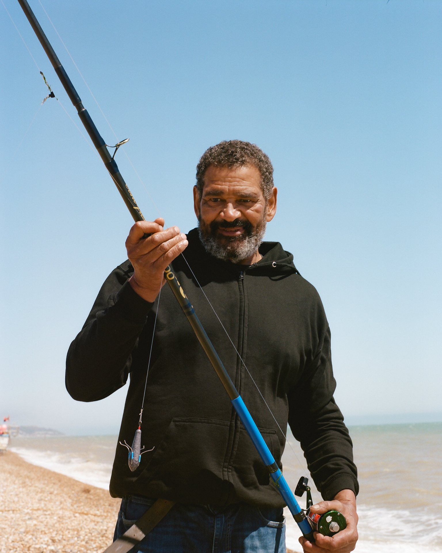 Man with grey beard in dark green hoodie holding fishing rod on beach with blue sky and sea in background.