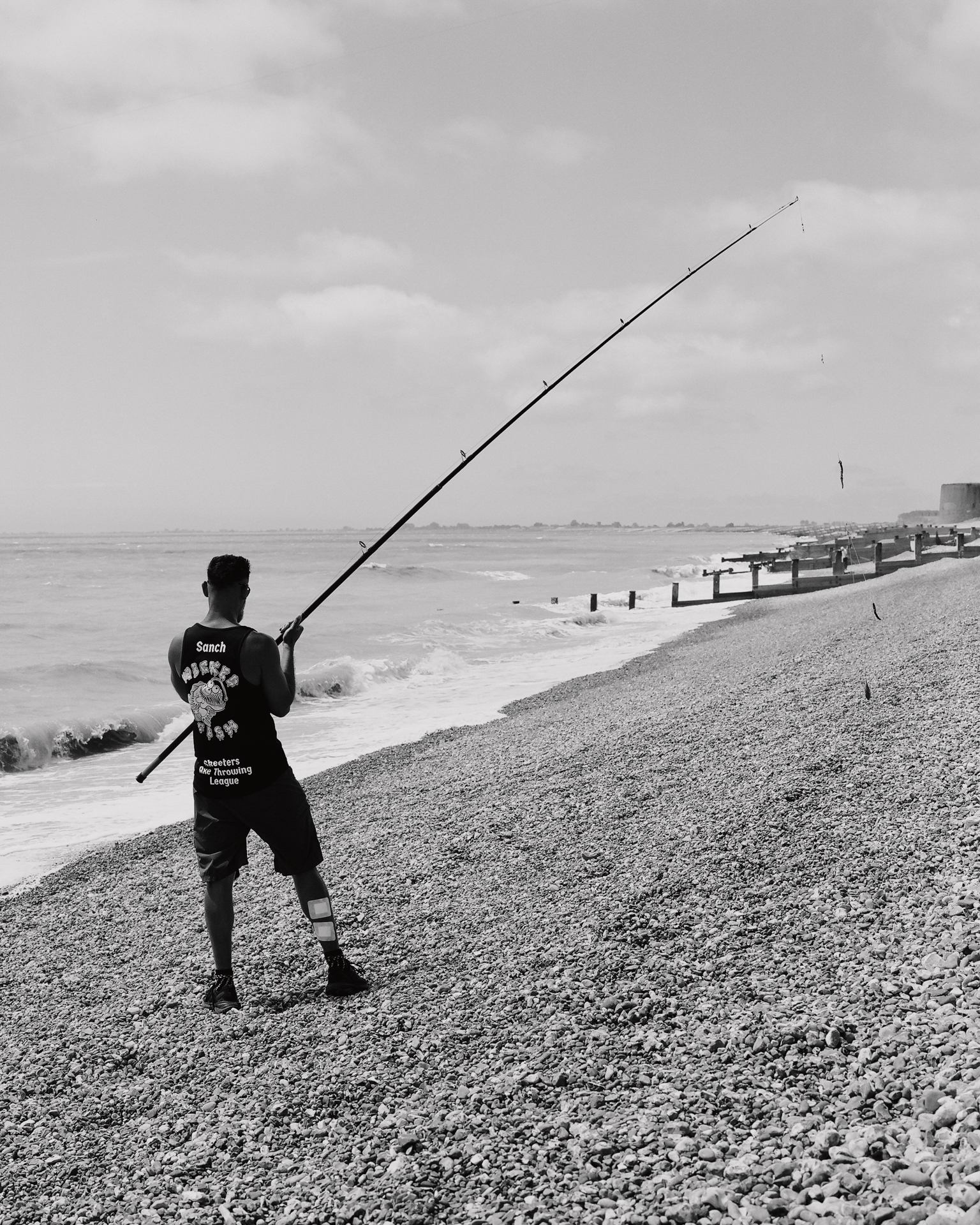 Person holding long fishing rod on pebble beach with sea, groyne, and pier structures visible under cloudy sky. Black and white image.