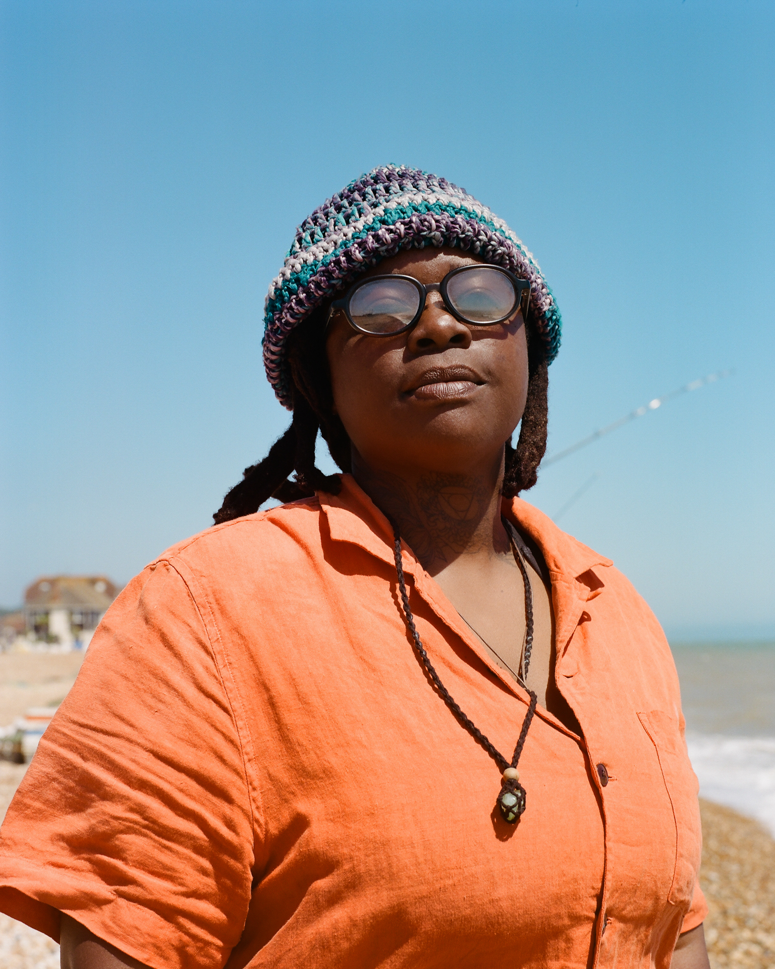 Woman wearing colourful knitted beanie and orange shirt with glasses, standing against blue sky background.