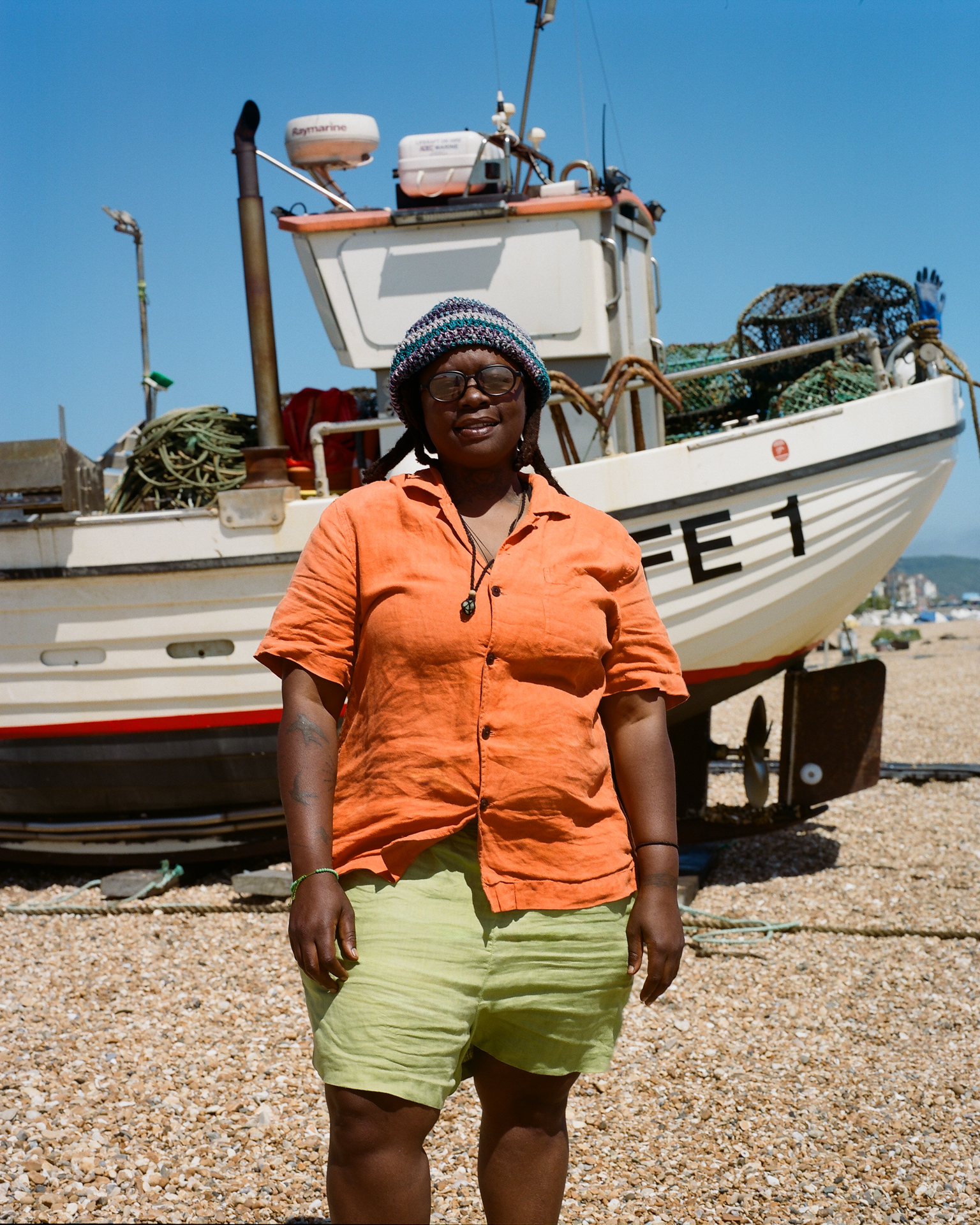 Woman in orange shirt and green shorts stands before white fishing boat with nets on beach under blue sky.