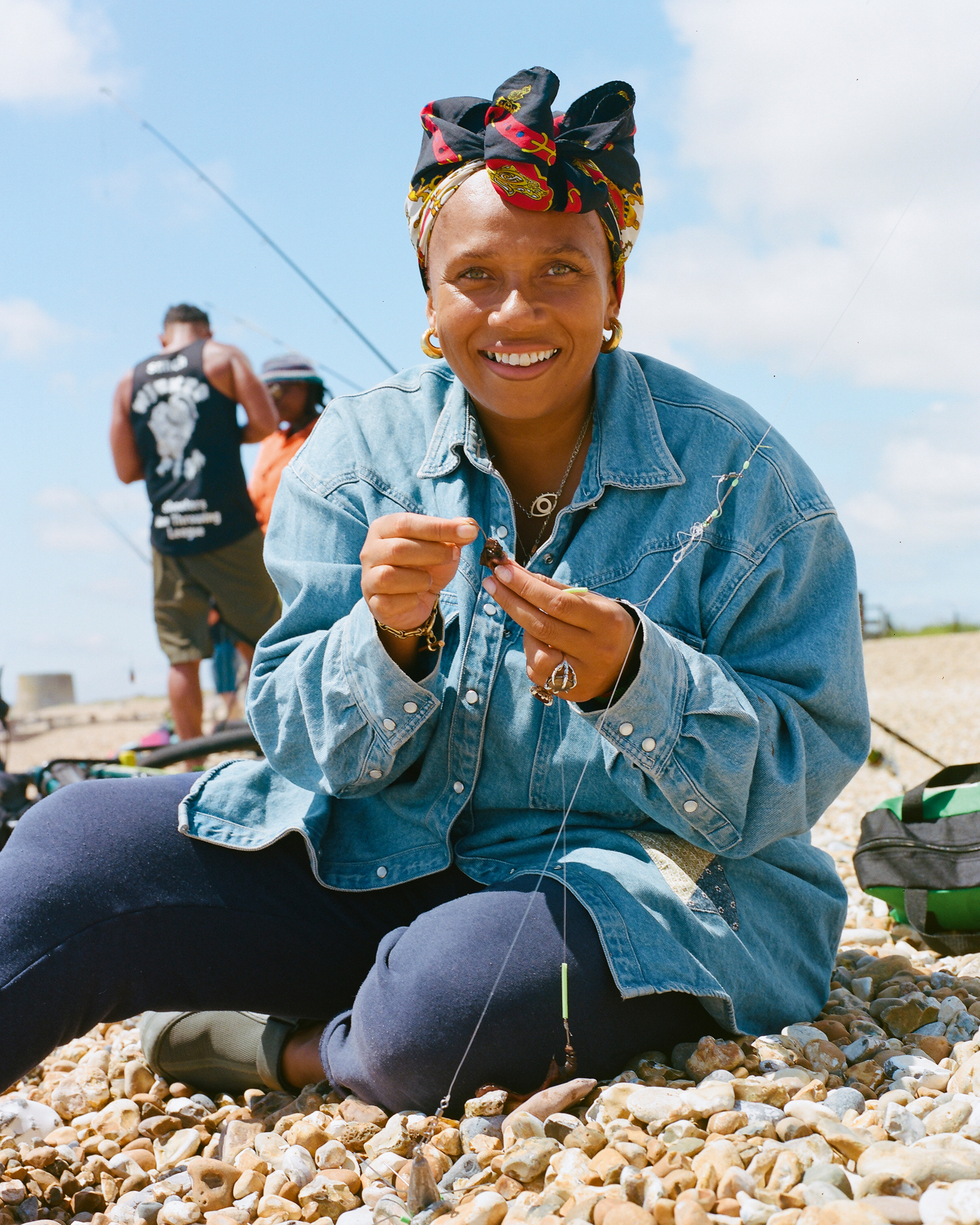 Smiling woman in denim jacket and colourful headscarf sits on pebble beach, holding small object. Man with fishing rod visible in background.