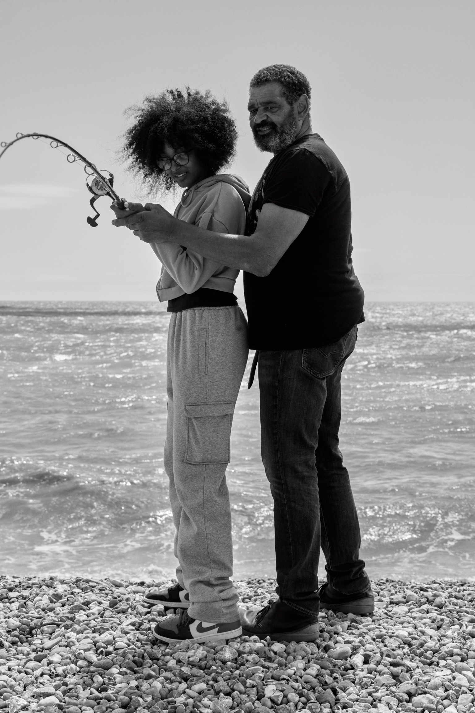 Black and white image of man and woman fishing together on pebble beach, both holding fishing rod, sea in background.