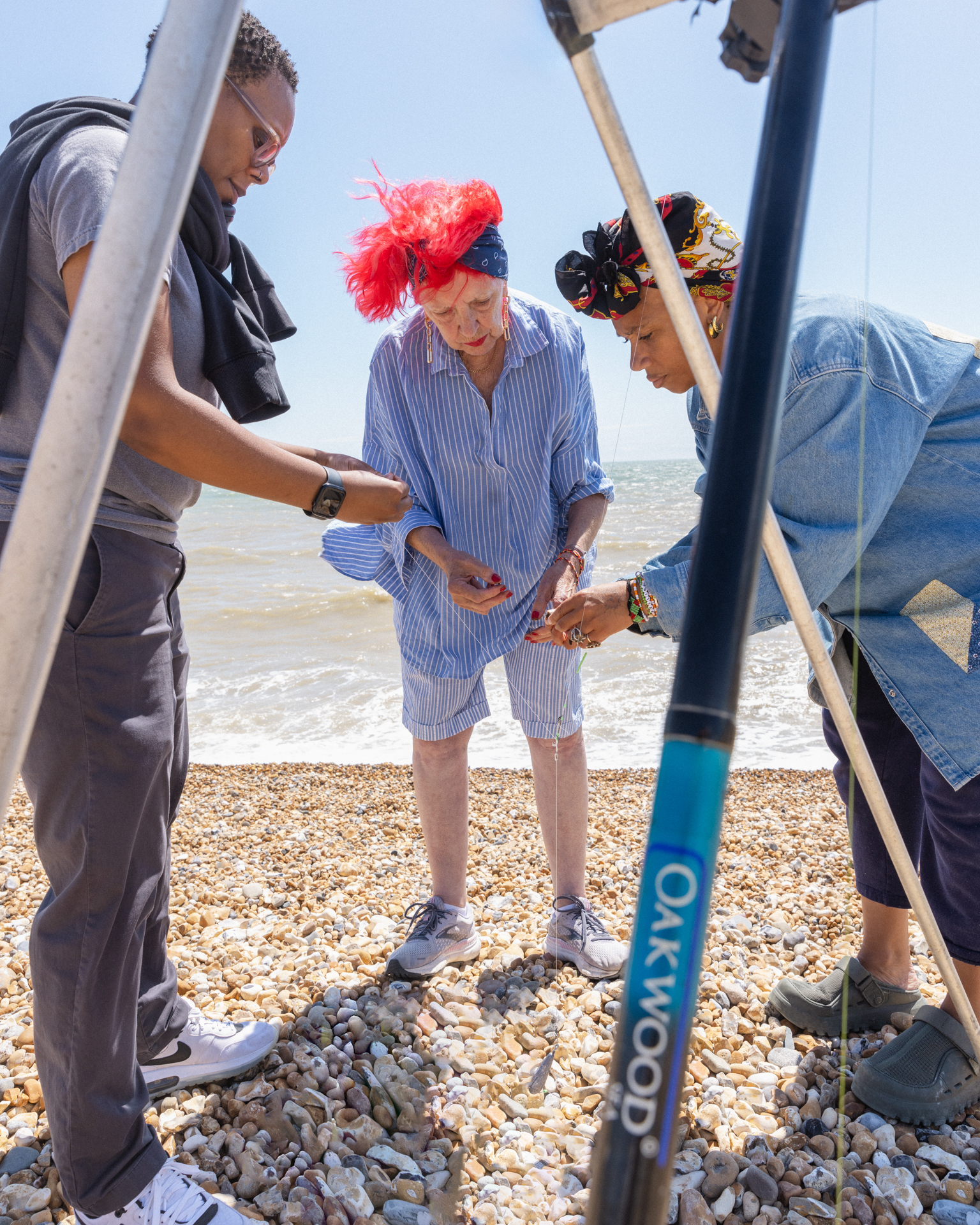 Two men assist elderly woman with red fascinator onto pebble beach using mobility aid frame near shoreline on sunny day.
